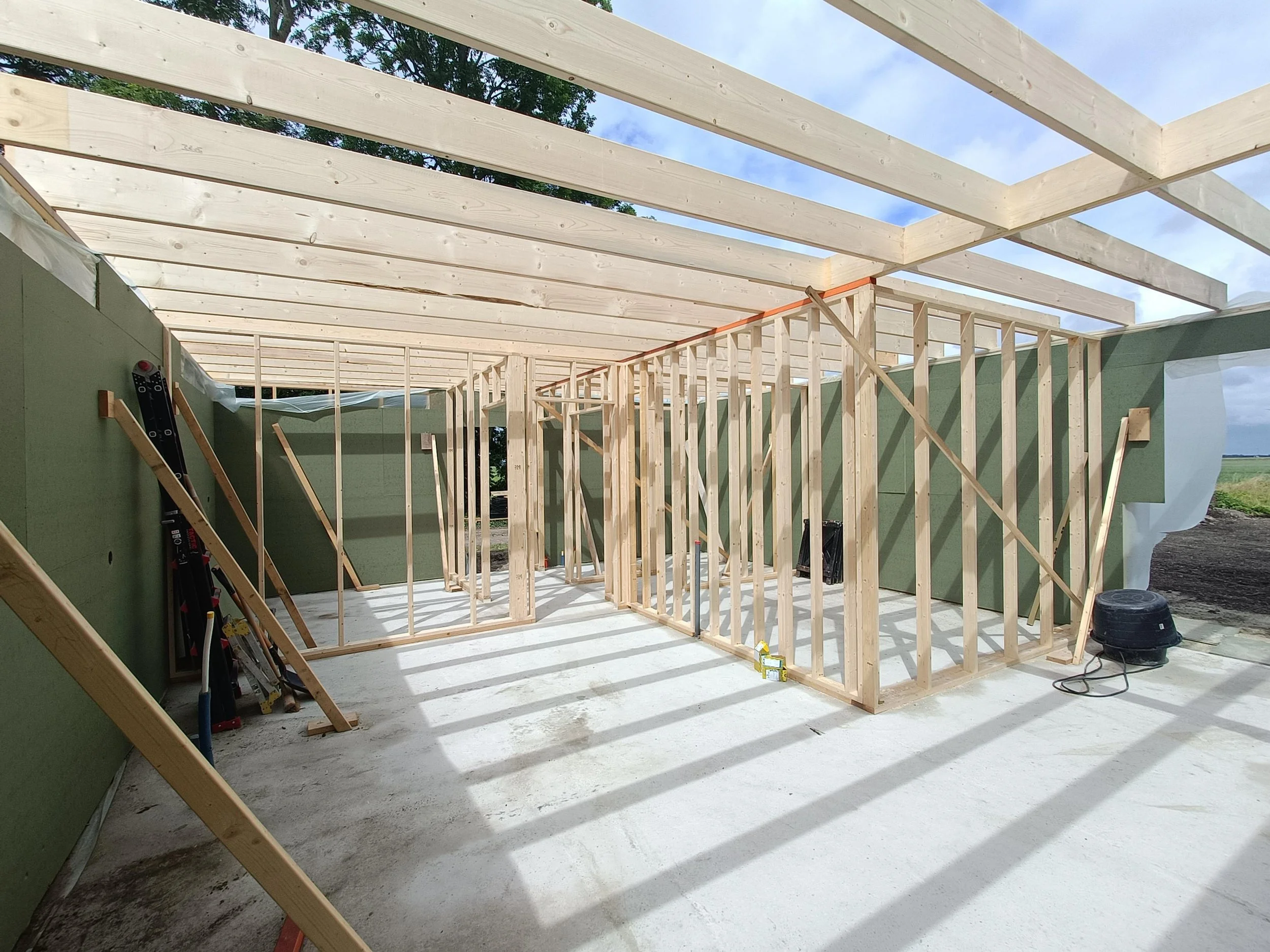 Construction site with wooden framing of a building under construction, green insulation panels on the walls, and a clear sky with some clouds overhead.