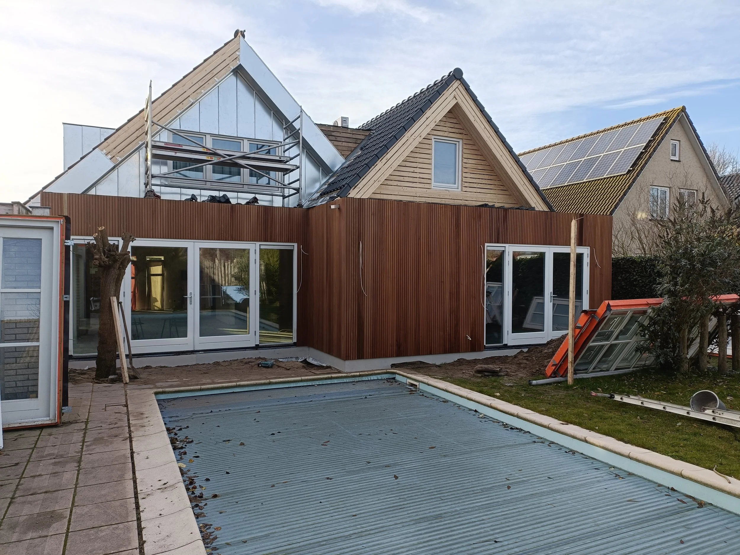 A house under construction with some parts recently renovated, including a new roof and solar panels. The backyard has a swimming pool with a cover, and construction tools and materials are scattered around.