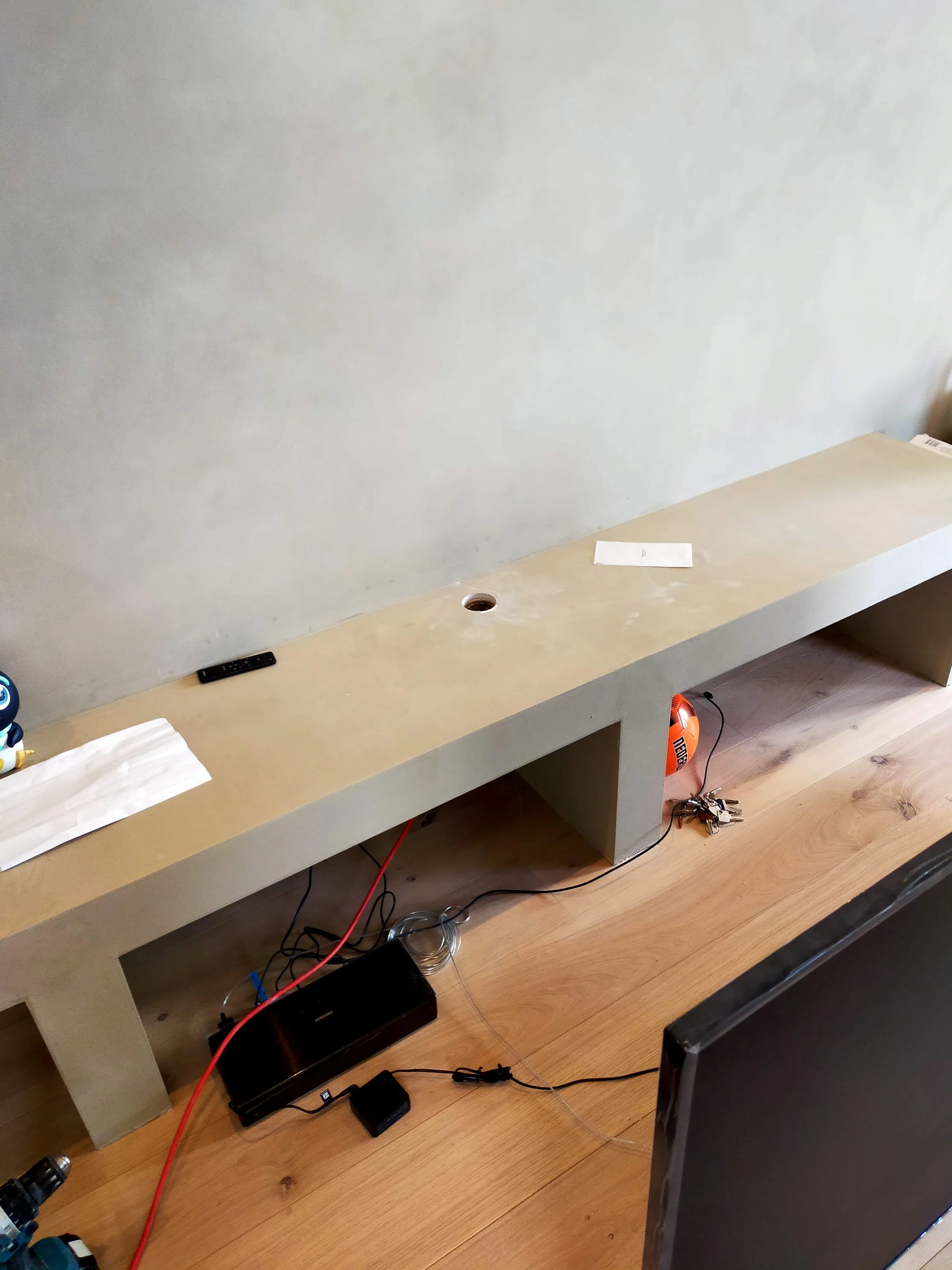 an unfinished wooden entertainment center with an open space in the center and openings on each side, placed against a light-colored wall, with various electronics and cords underneath and on top.