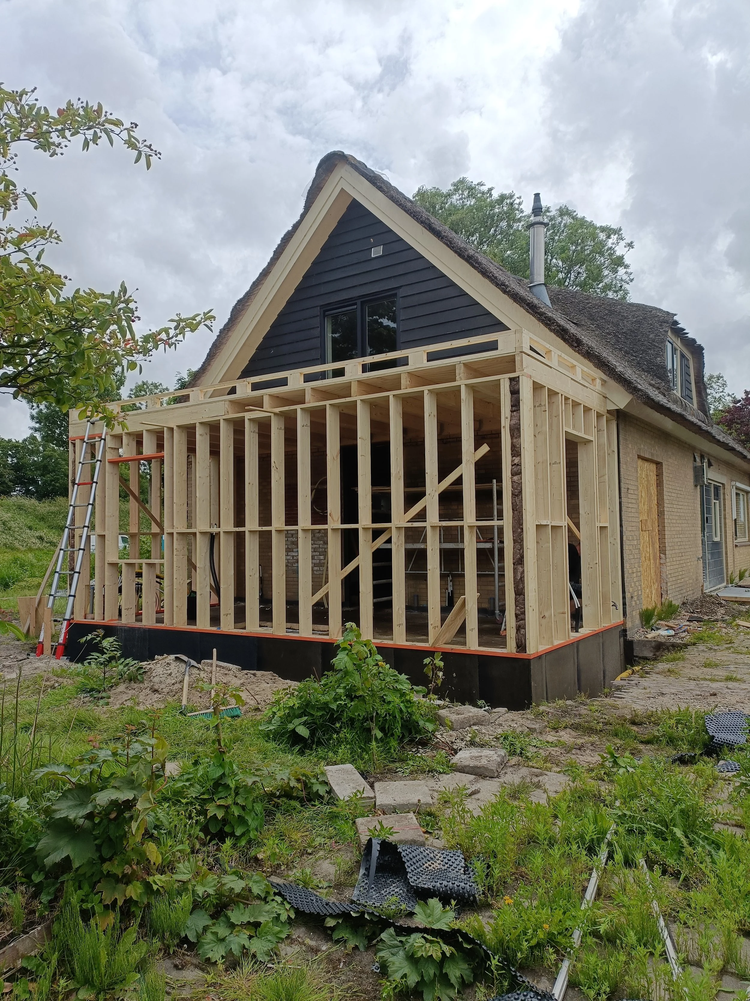 A house under construction with a wooden frame and black siding on the upper portion. The roof is steep and shingled, and there is a front porch area without finishing. The surrounding yard has green grass, plants, and construction materials, with a cloudy sky above.