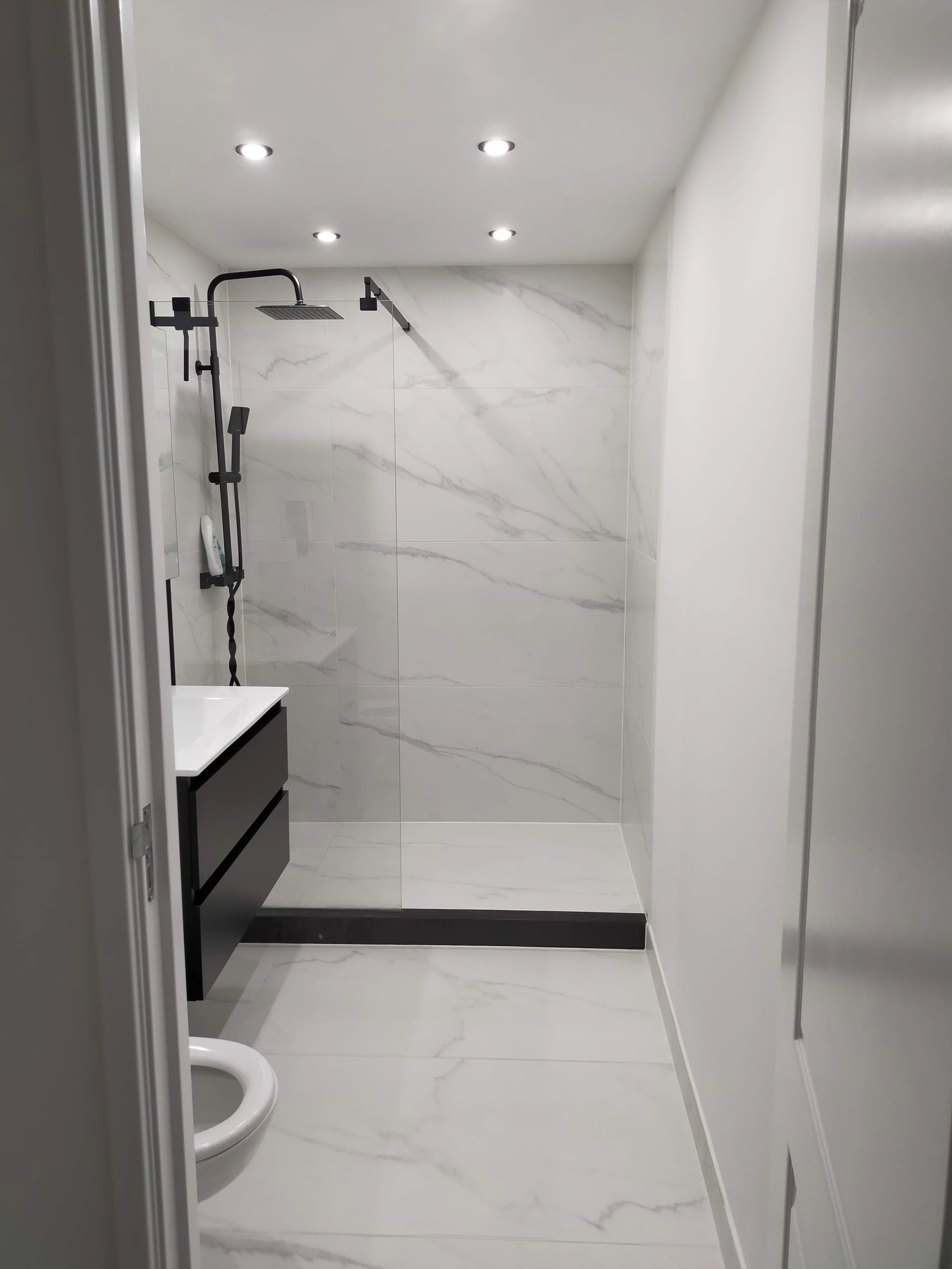 A modern minimalist bathroom with a walk-in shower enclosed by a glass panel. The shower has a rain shower head and black fixtures. The walls and floor are covered with white marble tiles with gray veins. There is a small vanity with a drawer on the left and recessed ceiling lights.