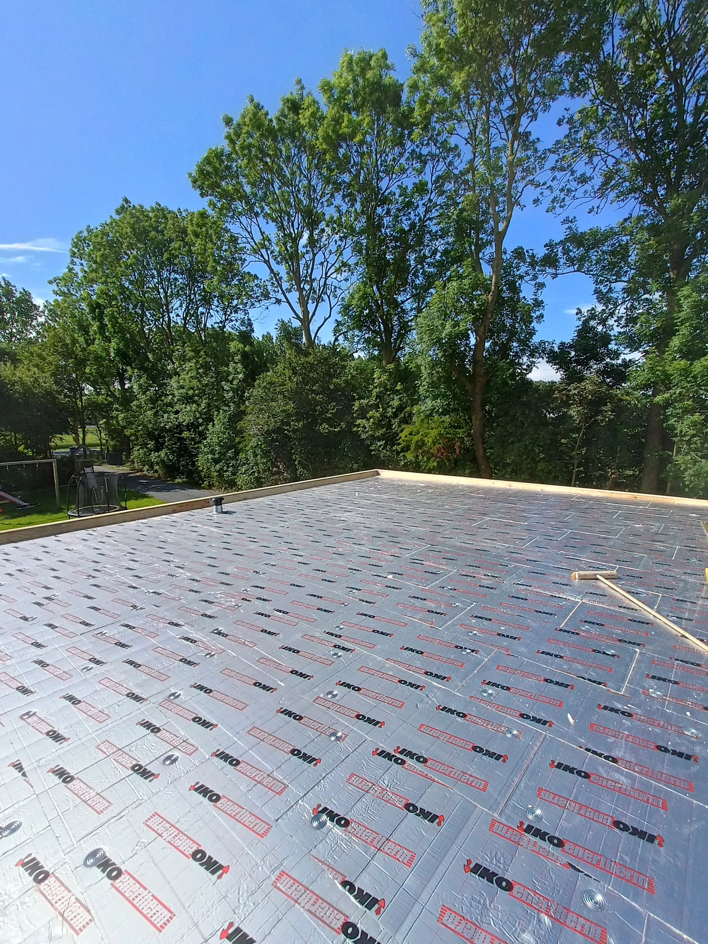 Flat roof with insulation panels and fasteners, surrounded by trees under a clear blue sky.
