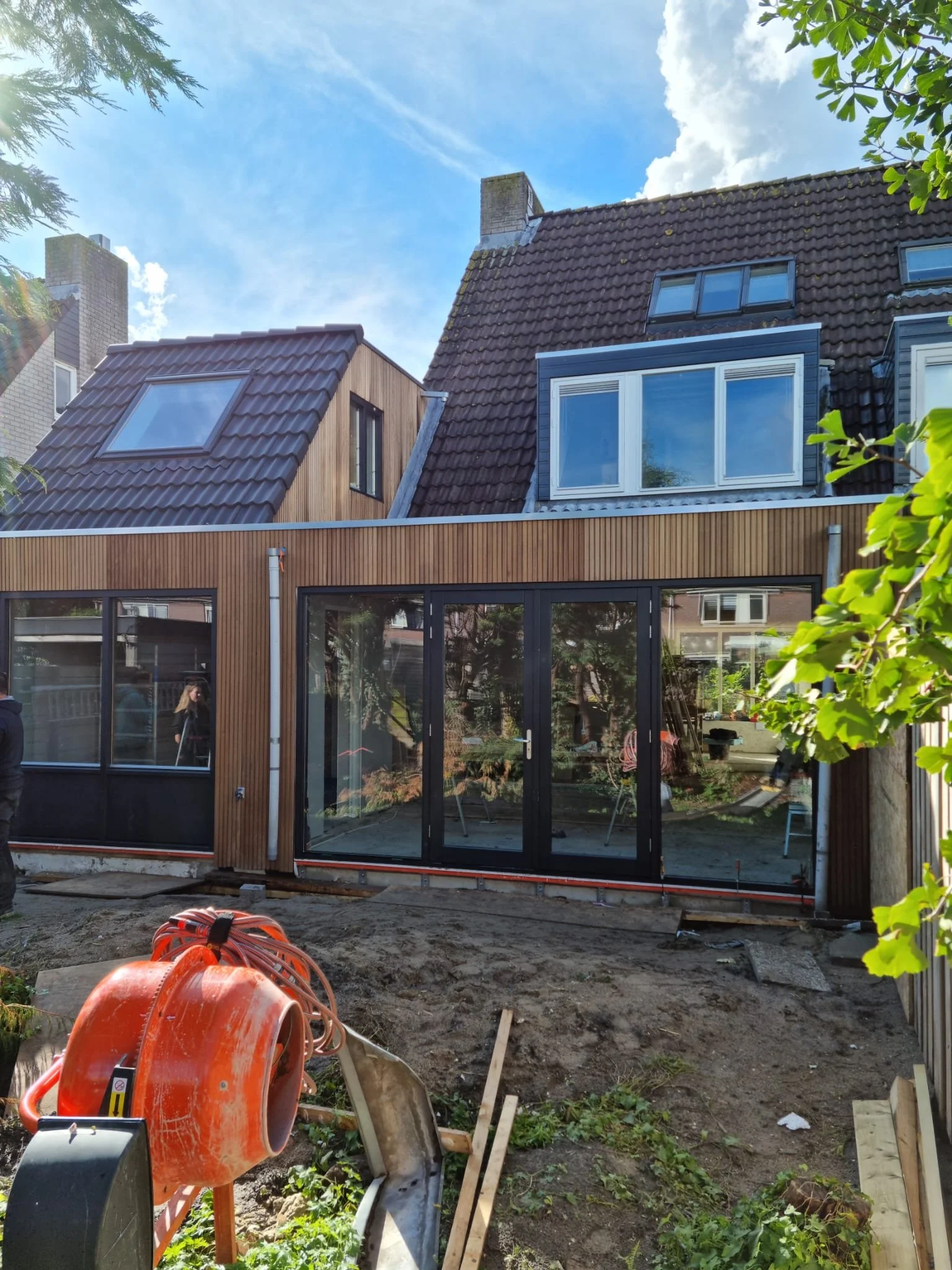 Construction site in backyard with a partially built sunroom with glass sliding doors, construction tools, and a concrete mixer, with residential houses and trees in the background.