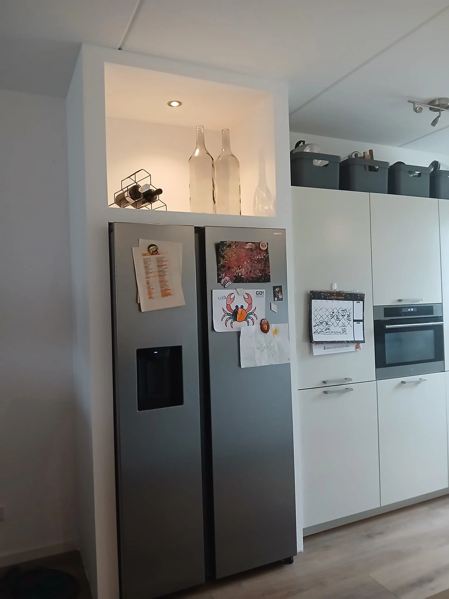 Kitchen with a stainless steel refrigerator, magnetic calendar, and notes on a door. White cabinet with black storage boxes on top and a shelving nook with decorative bottles and wine rack.