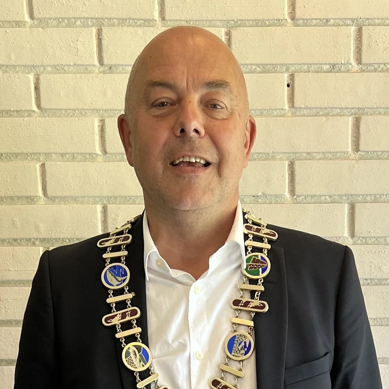 En mann med blanke frisyr, iført mørk dress og hvitt skjorte, smiler foran en lys murvegg, bærer en lang kjede med diverse medaljer og symboler.