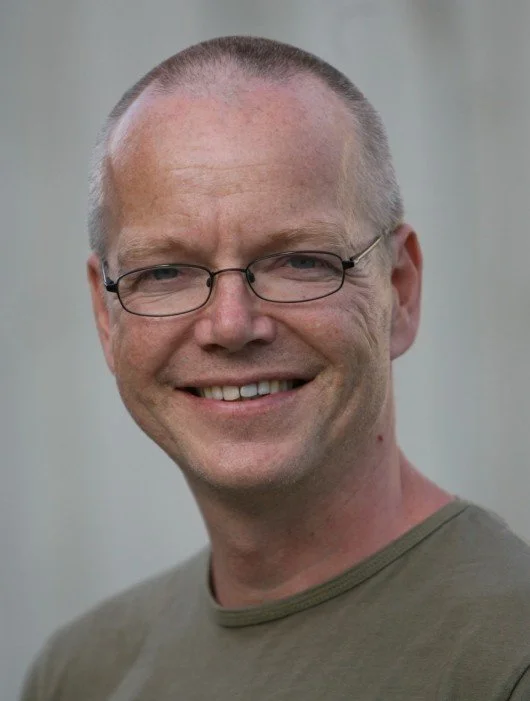 Portrett av en smilende mann med briller, lyst hår, og en grå t-skjorte, mot en nøytral bakgrunn.