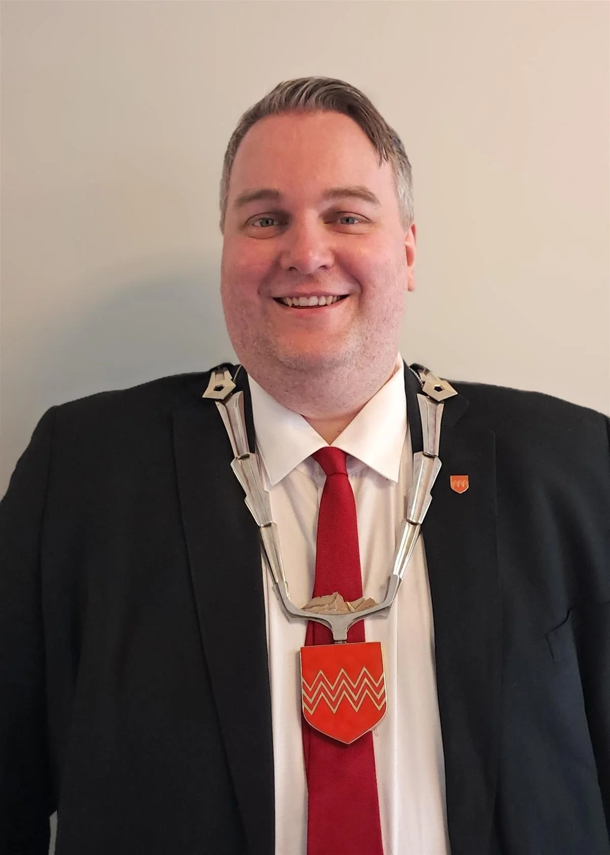 En smilende mann i svart dress, hvit skjorte og rød slips, iført en medalje med et skjold i gul og rød farge rundt halsen.
