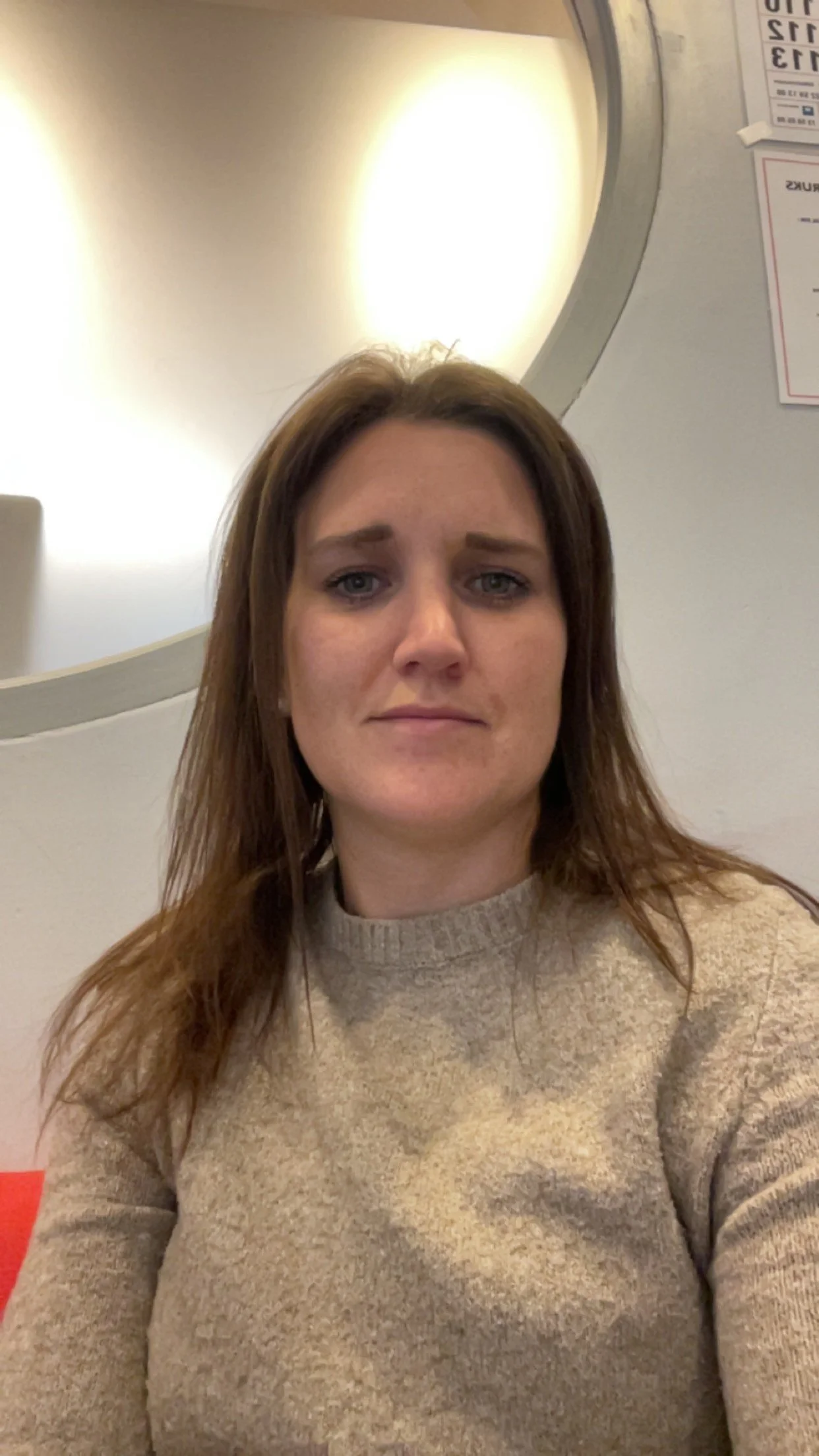 Et bilde av en kvinne med langt brunt hår som tar en selfie. Hun har på seg en lys beige genser og står innendørs, med en rund speil og en veivisningstavle på veggen bak henne.