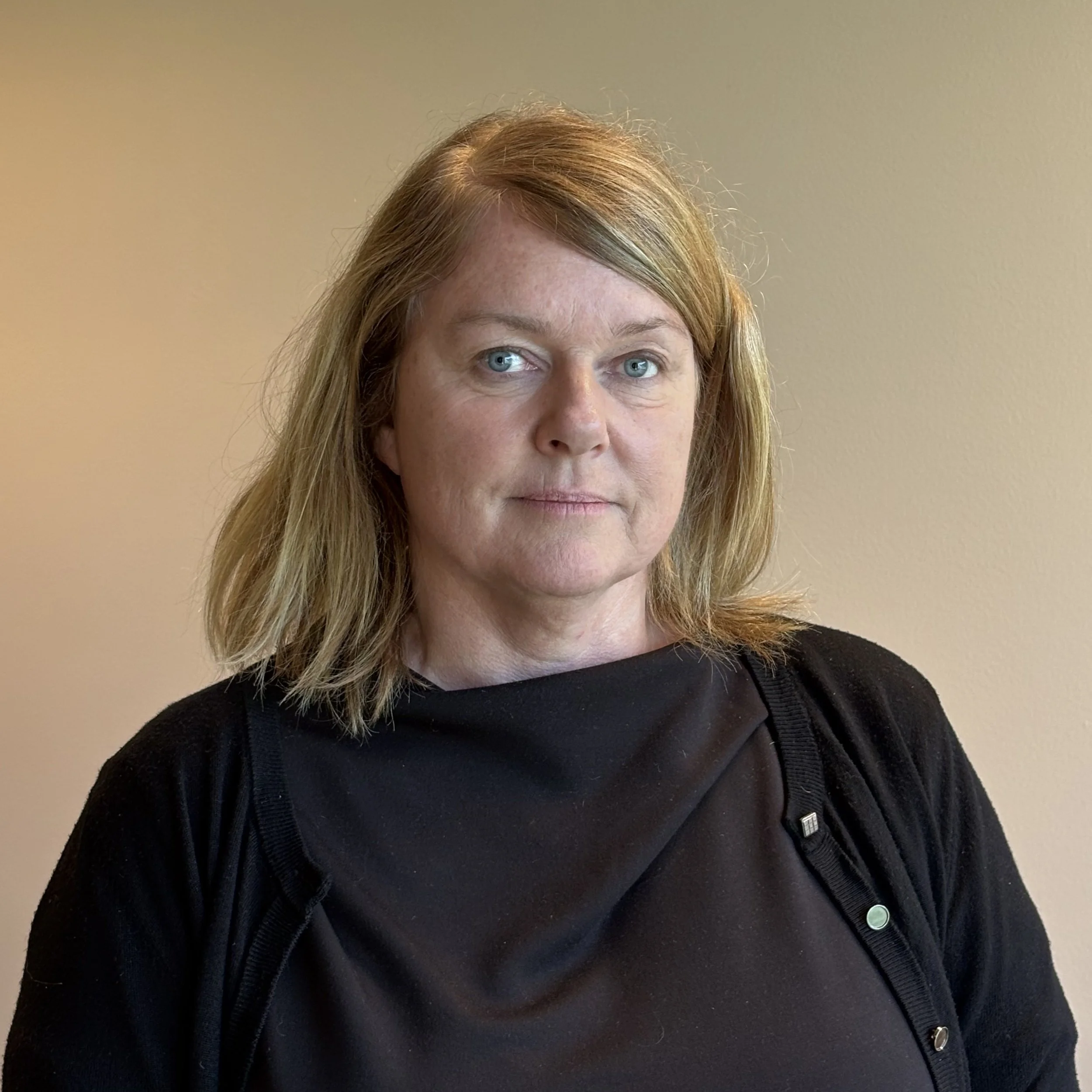 Nærbilde av en kvinne med blond hår og blå øyne, iført svart genser og jakke, foran en beige vegg.