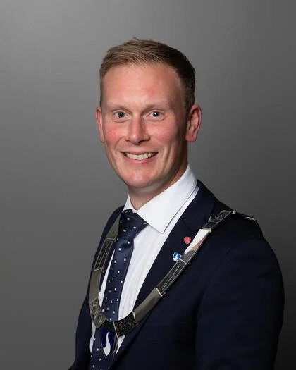 En mann i formell dress med blå dress og hvit skjorte, smilende, på et grå bakgrunn, iført et belte rundt halsen.