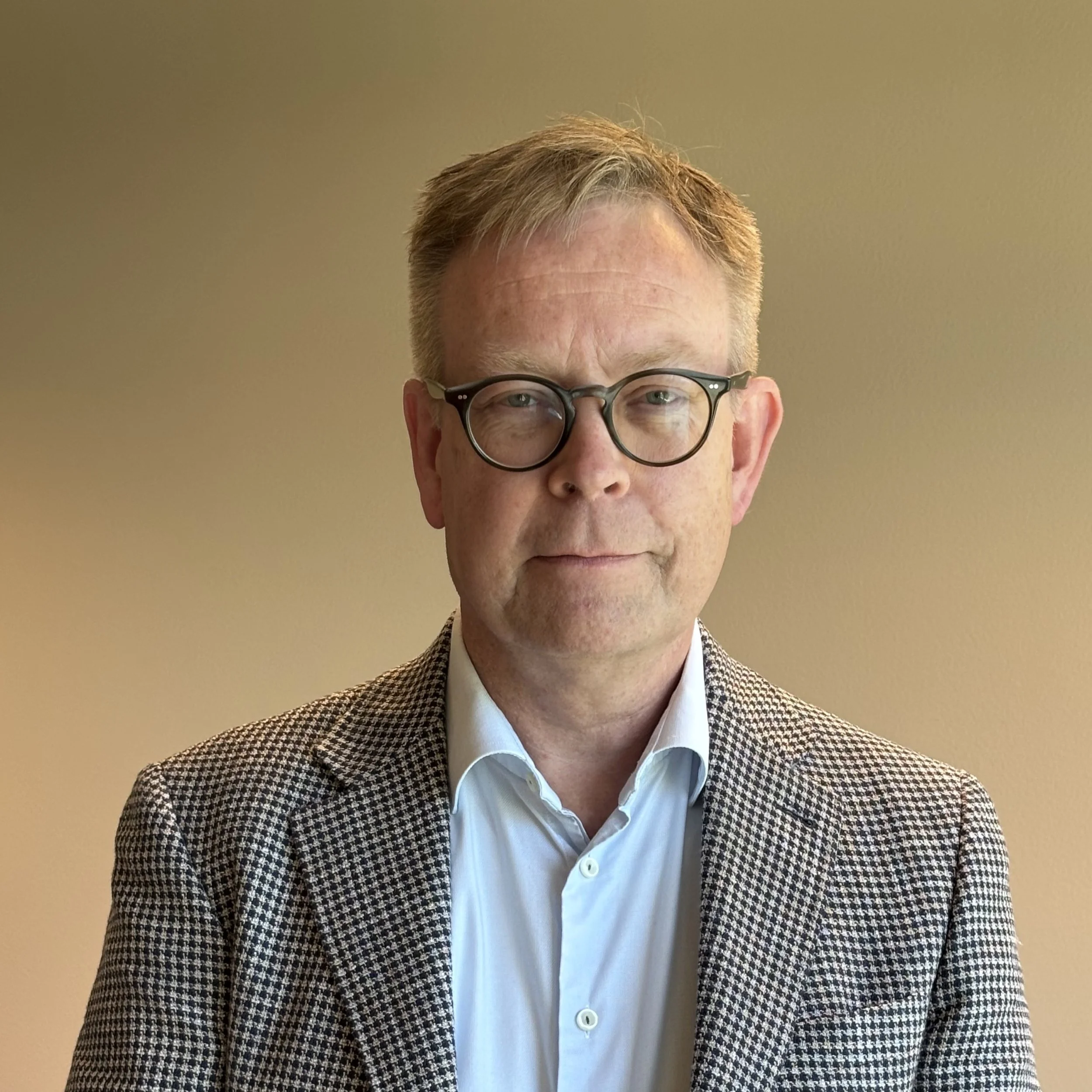 Et nærbilde av en mann med briller, iført en blazer og hvit skjorte, mot en beige bakgrunn.
