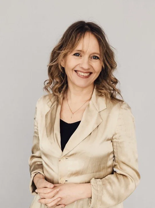 Kvinne som smiler, har brunt, krøllet hår og bærer en beige blazer over en svart topp.