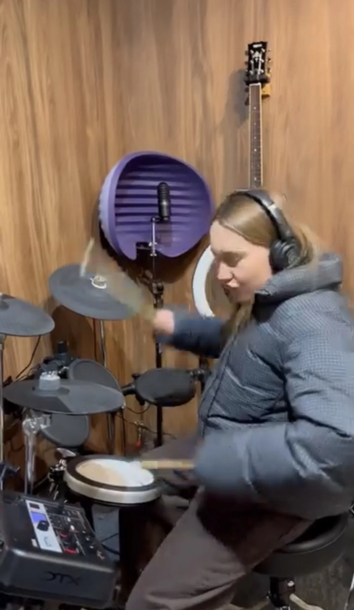 Une femme joue de la batterie dans une pièce avec un mur en bois. Elle porte un casque. Une guitare est accrochée au mur en arrière-plan.