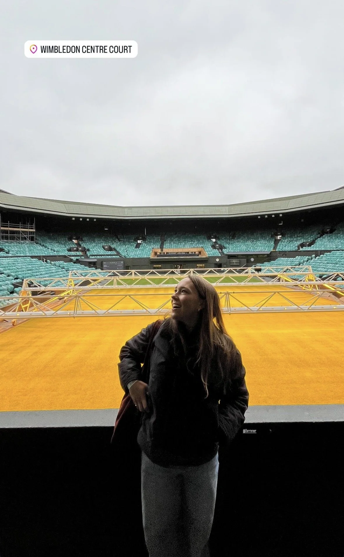 Une femme se tenant devant la scène vide d'un stade de tennis en plein air, avec des sièges vides et une structure métallique orange au premier plan, à Wimbledon.