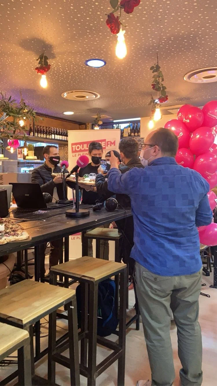 Groupe de personnes lors d'une interview ou d'une séance de radios dans un restaurant, avec des microphones, ballons roses, et décor floral sur le plafond.