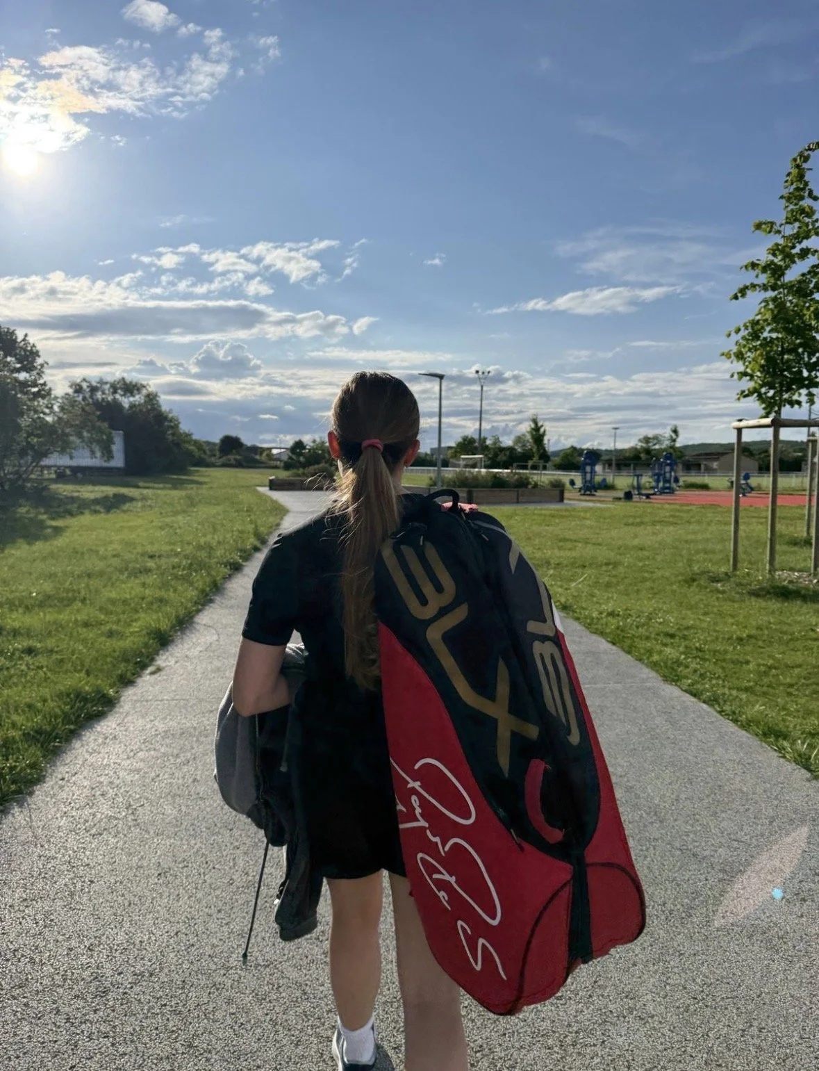 Fille avec un sac de tennis marche sur un sentier dans un parc en plein air, avec une aire de jeux en arrière-plan, sous un ciel partiellement nuageux.