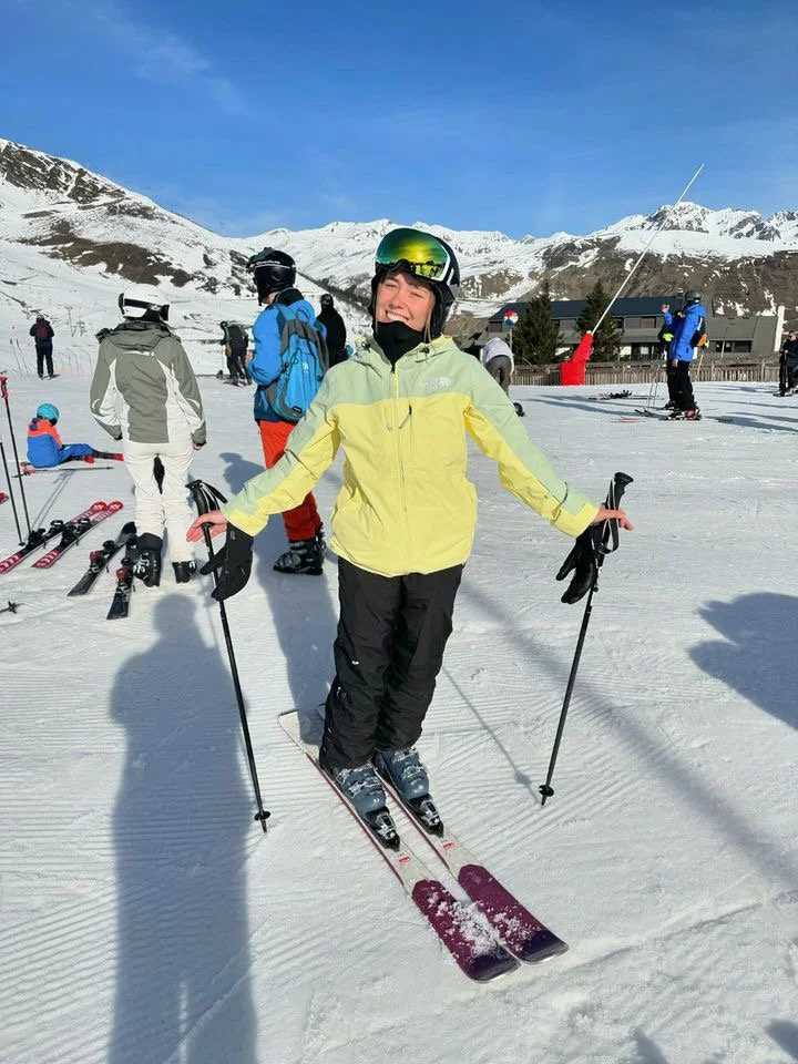 Une jeune femme souriante en ski sur une piste enneigée, entourée d'autres skieurs et montagnes en arrière-plan.