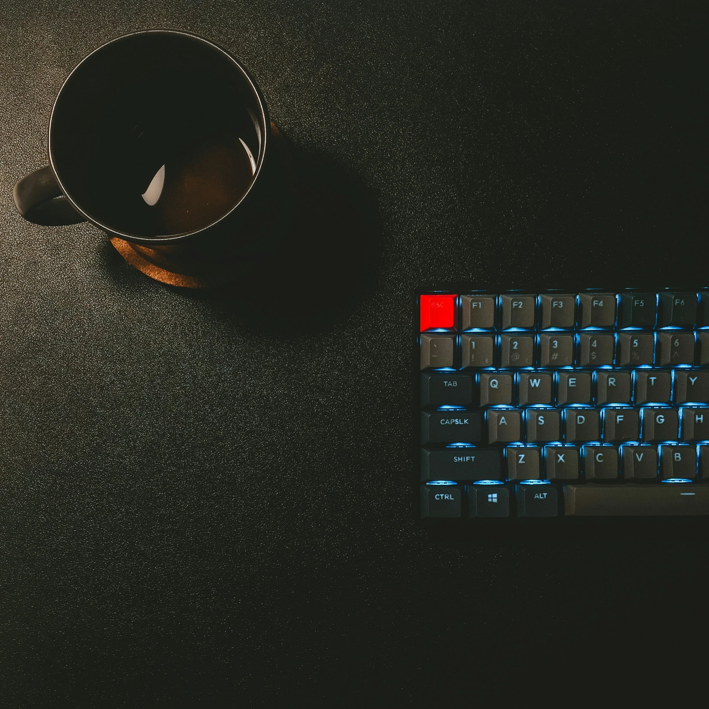 Top-down view of a black coffee mug placed on a dark speckled desk next to a computer keyboard with illuminated keys.