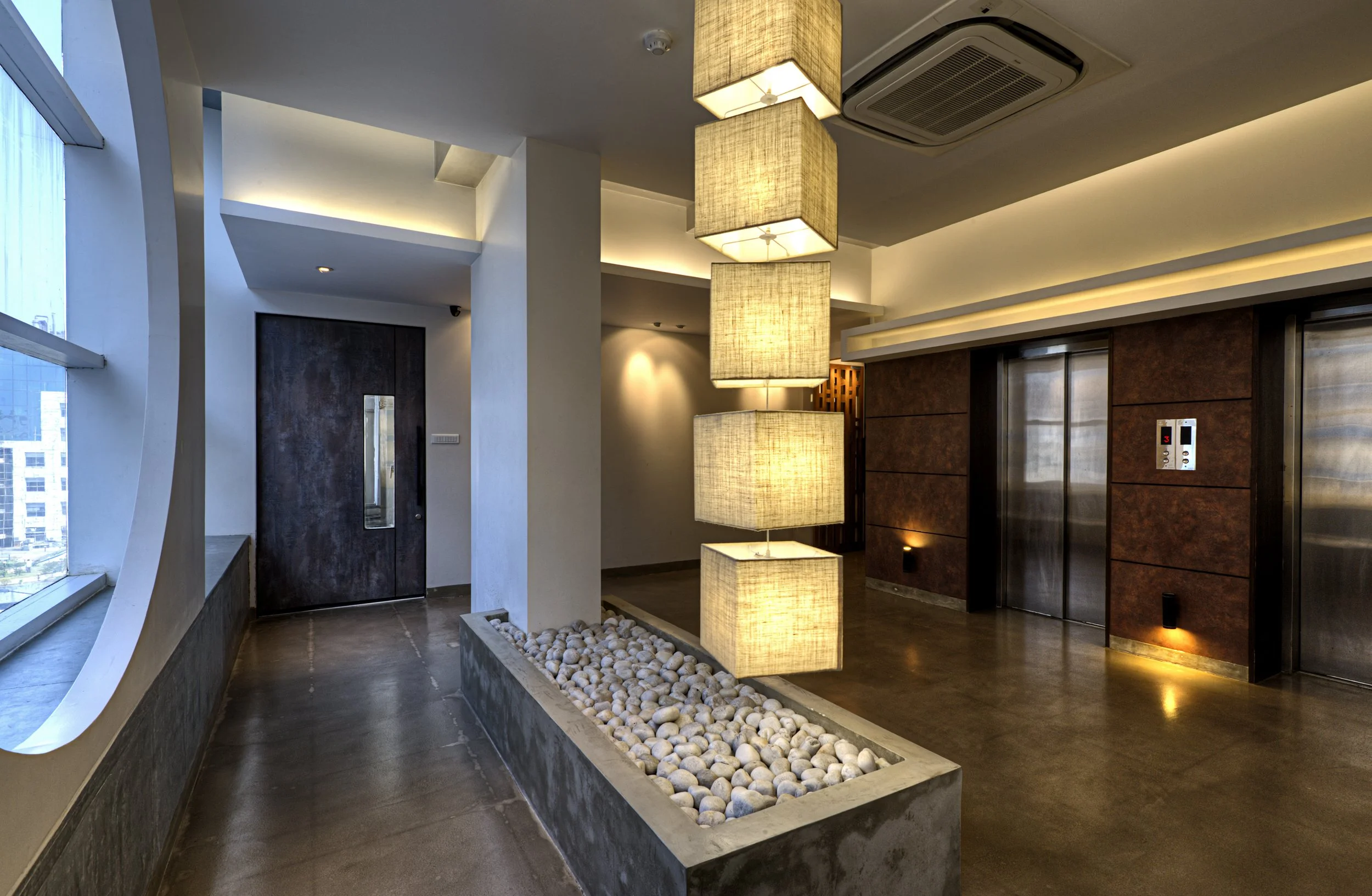 Modern hotel lobby with a large window, white walls, and warm lighting. Features a decorative light fixture with multiple rectangular fabric shades and an elevator with wooden paneling.