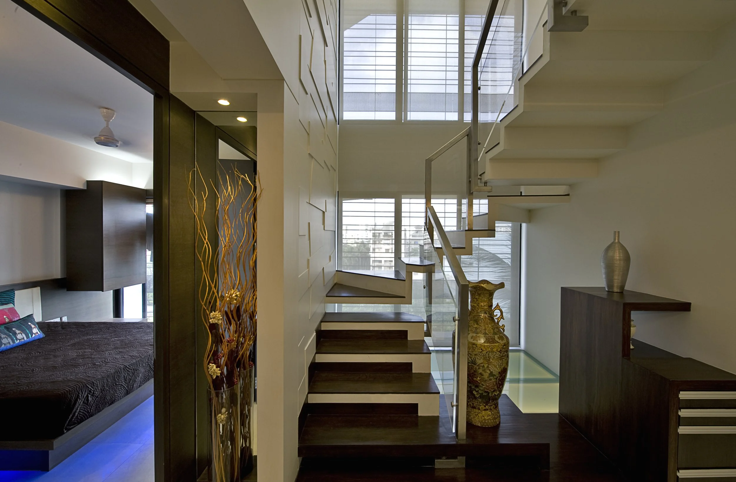 Interior view of a modern home with staircase, large windows, decorative vases, and a sitting area with a bed, pillows, and window blinds.