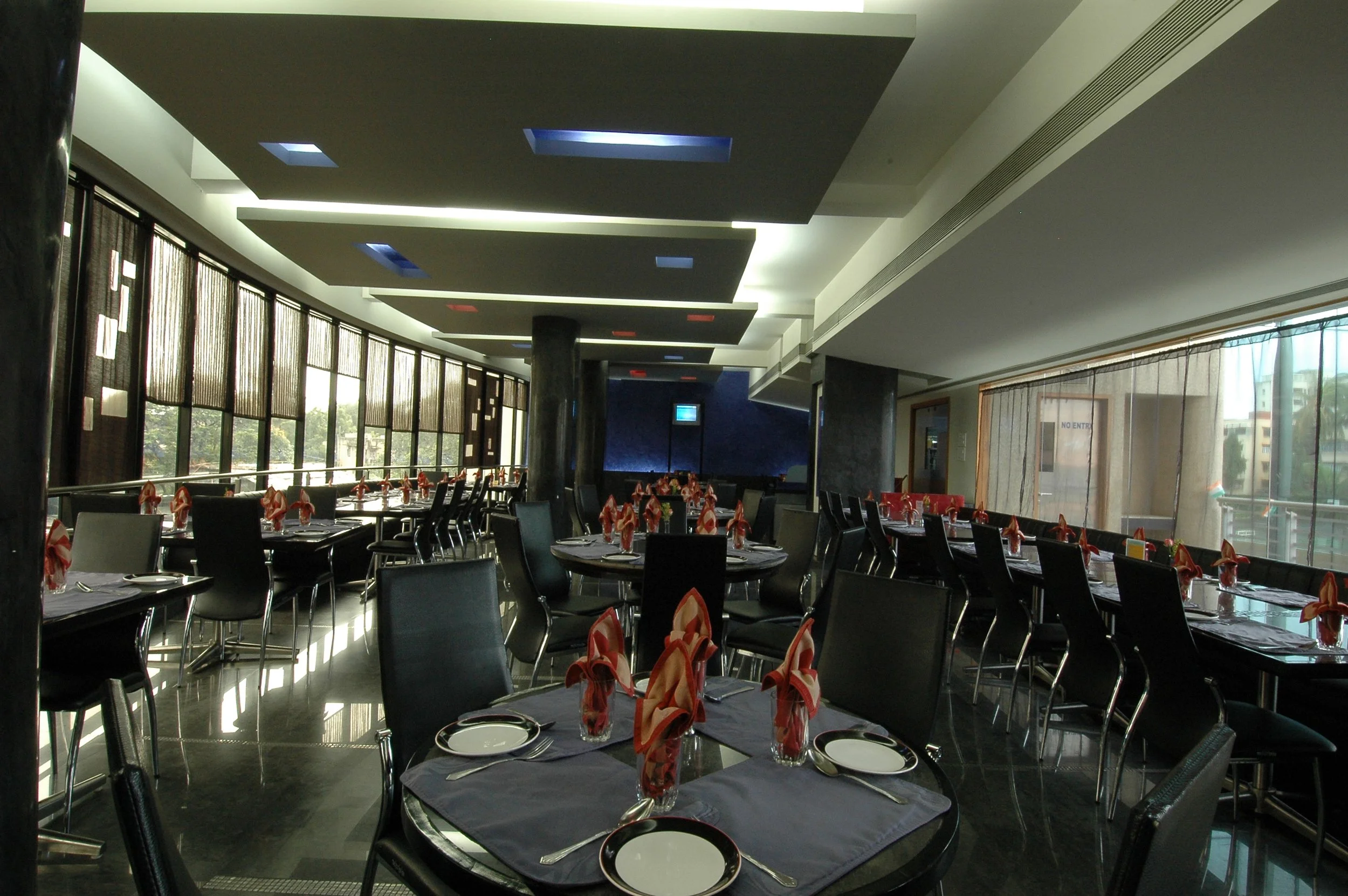 Interior of a modern restaurant with large windows, black chairs, tables set with plates, silverware, and folded napkins, and a ceiling with geometric black panels and lighting.