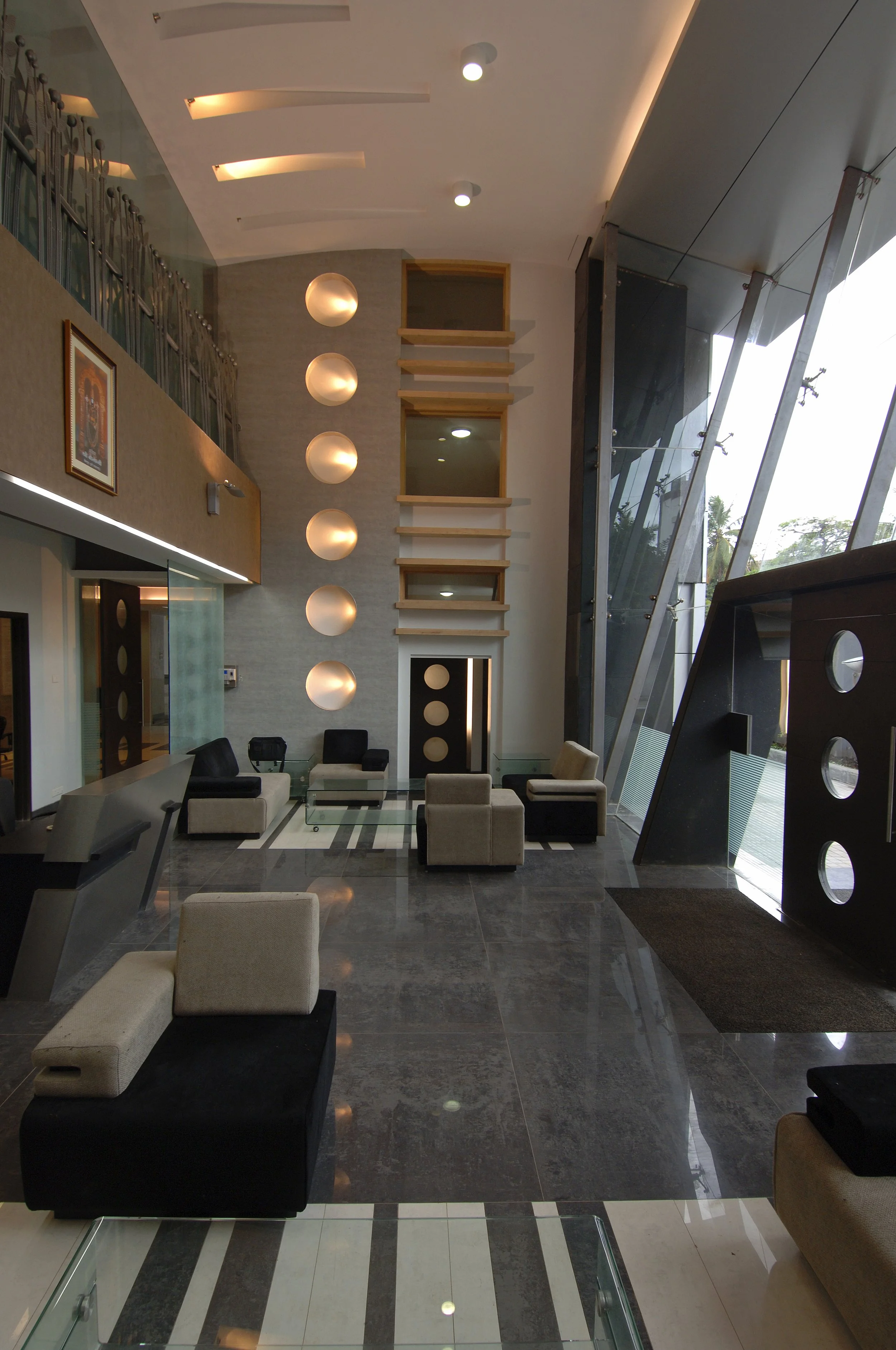 A modern hotel lobby with a high ceiling, illuminated spherical hanging lights, a seating area with black and beige chairs, and a large glass wall with an angled metal framework on the right side. SVKM girls hostel.