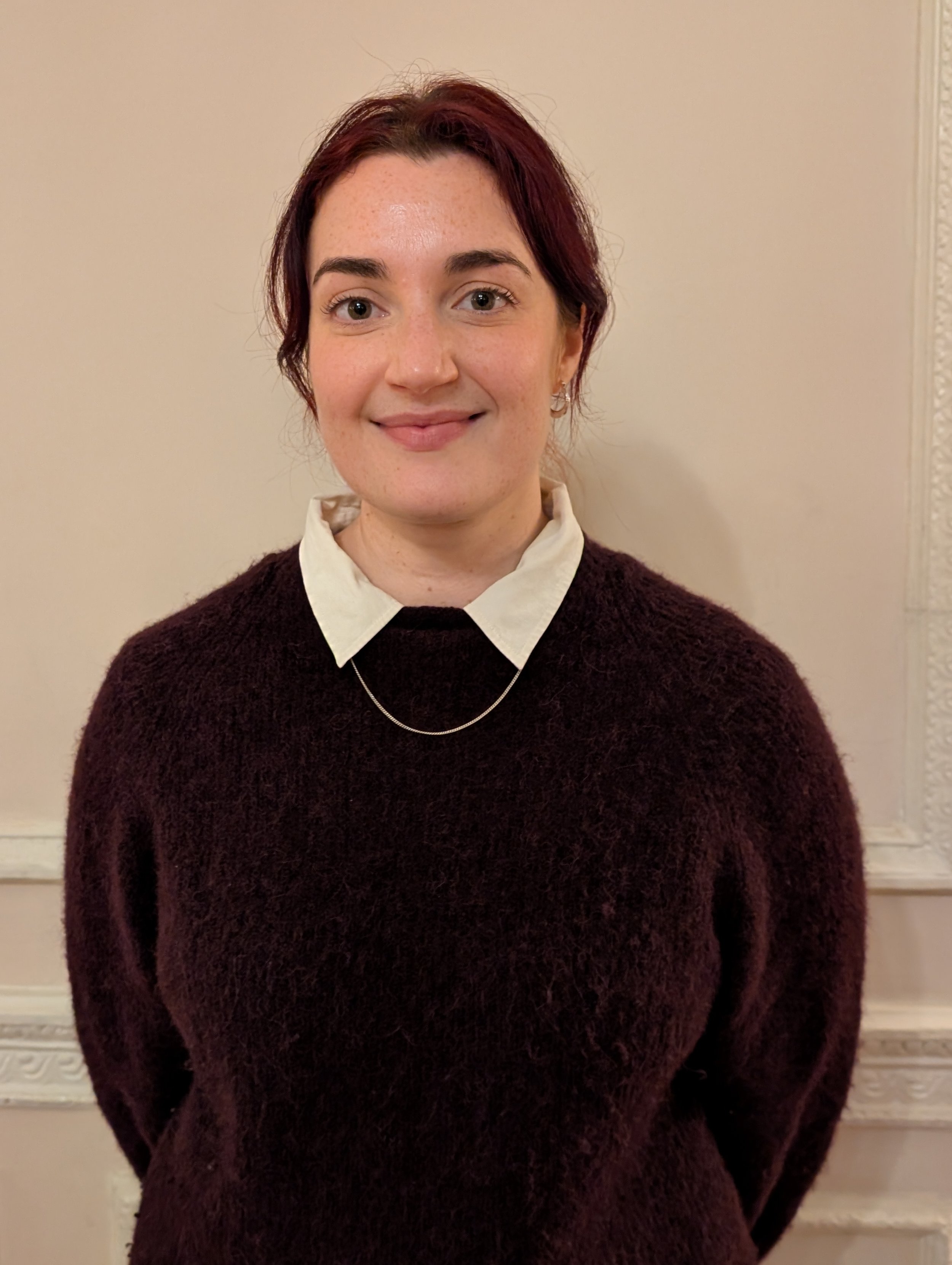 Anouk stands smiling in a burgundy jumper with a cream collared shirt and silver necklace against a white wall with some cornicing.