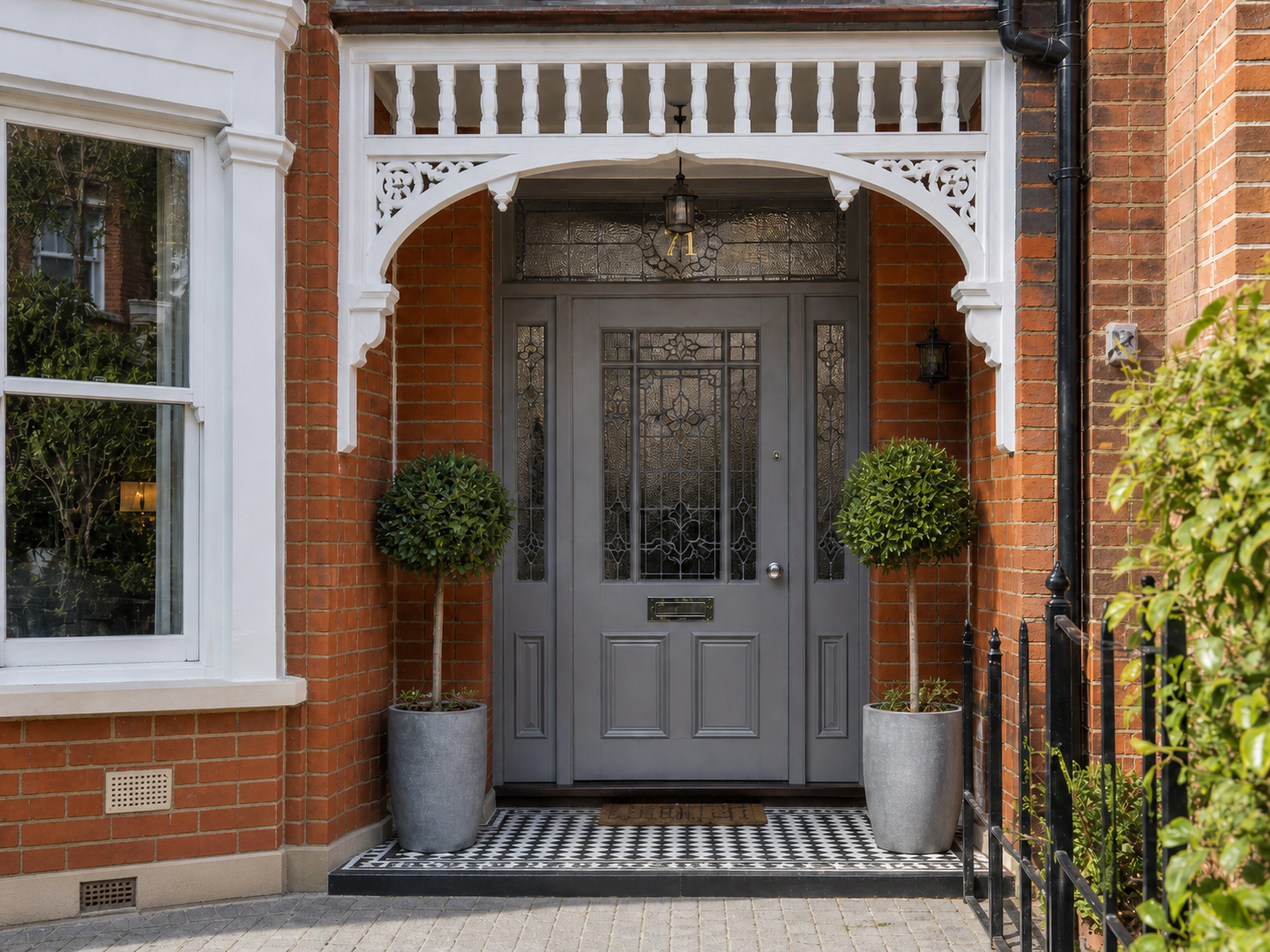 A gray front door with decorative glass panels, flanked by two potted topiary trees in gray planters, on a patterned tile porch with a black iron railing, brick walls, a window to the left, and a pergola with white decorative woodwork above.