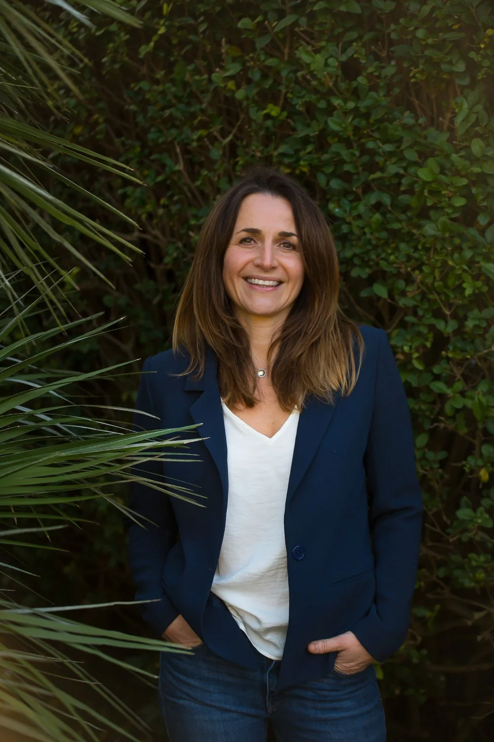 A woman with shoulder-length brown hair, wearing a dark blue blazer and white shirt, smiling and standing with her hands in her pockets outdoors in front of green foliage and plants.