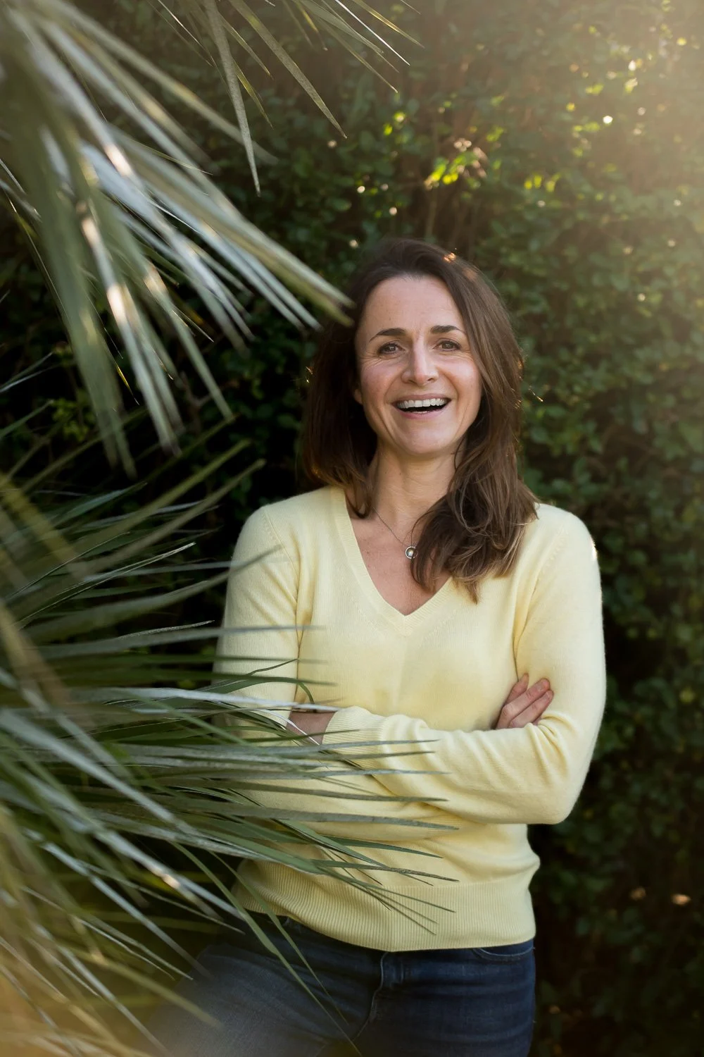 A woman with medium-length brown hair, smiling, standing outdoors among green plants and bushes, wearing a yellow sweater, with sunlight filtering through the trees.
