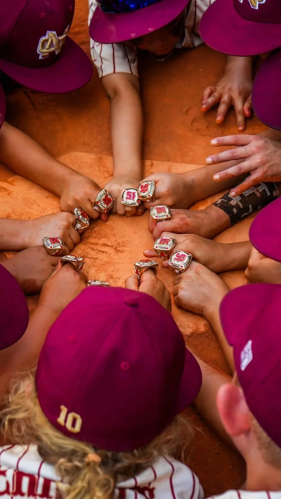 Young baseball players in maroon caps and jerseys, sitting on the field, showing off their championship rings.