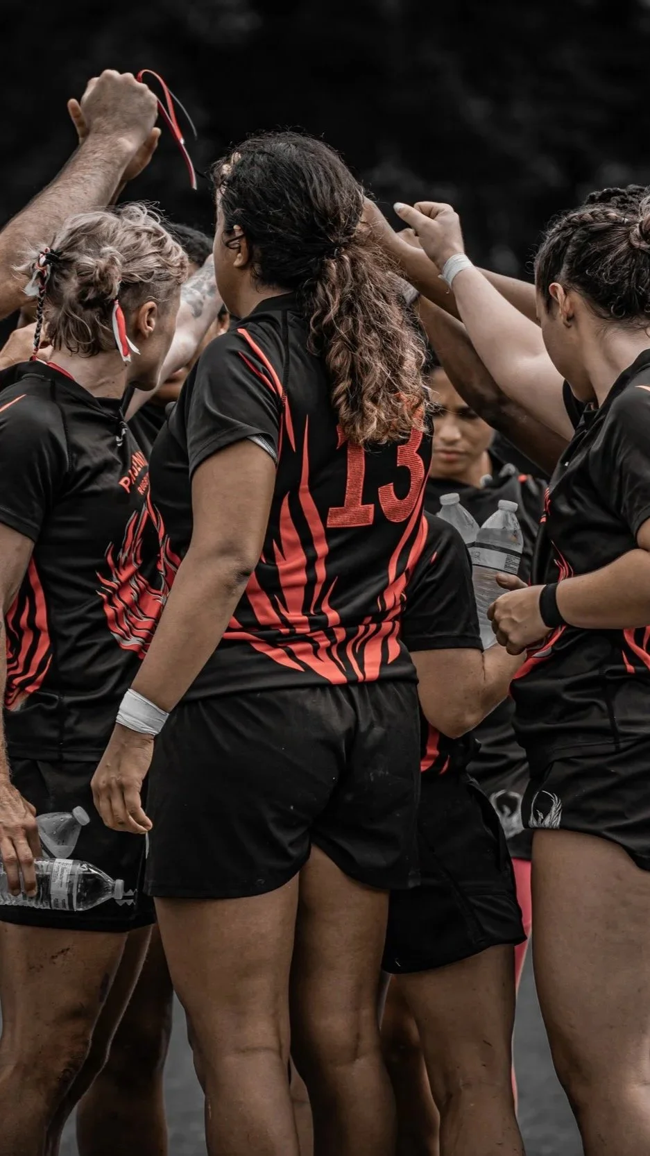 Women’s rugby team huddled together during a game, wearing black and red uniforms, some holding water bottles.