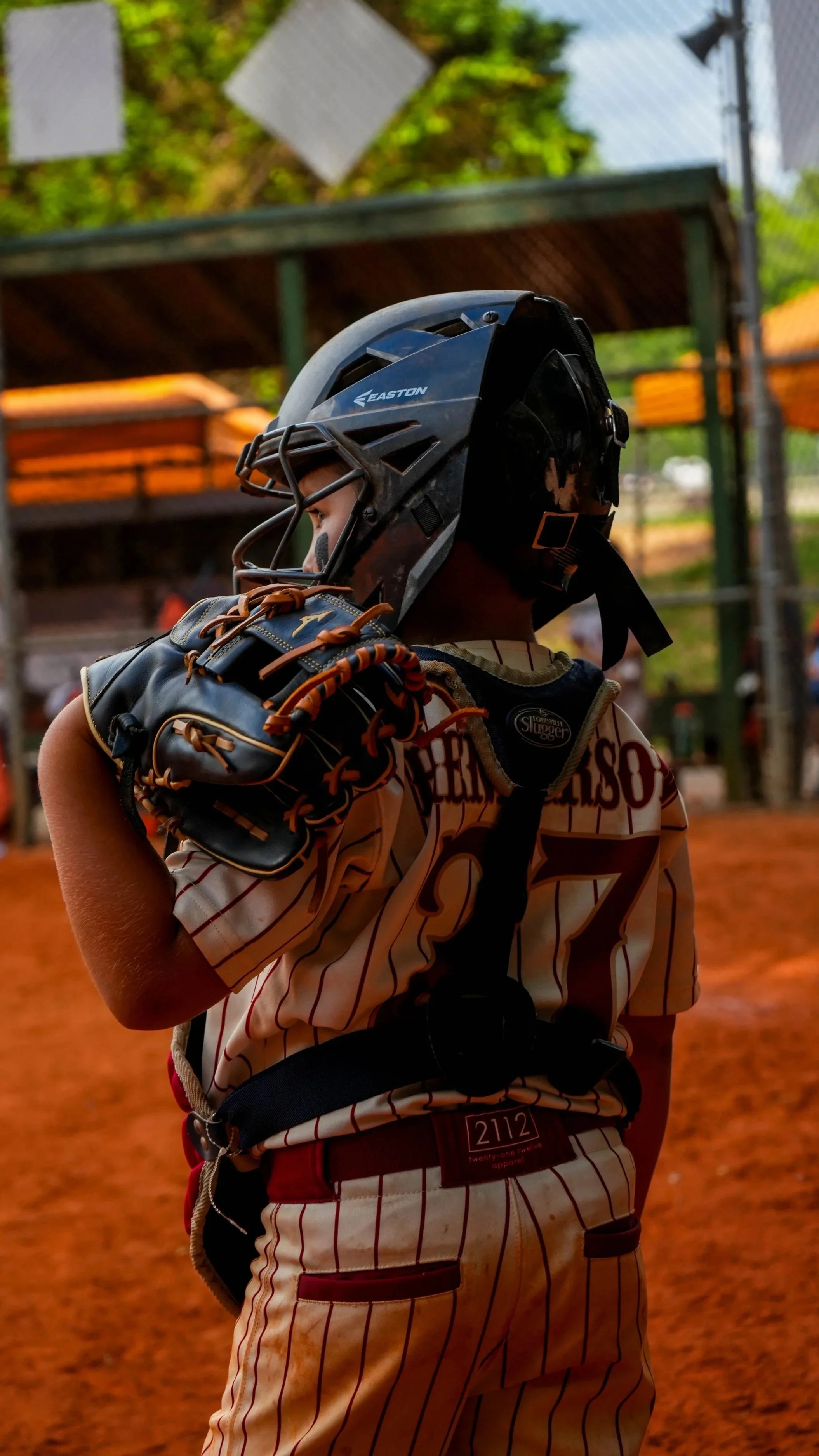 A young baseball player wearing a black helmet, striped jersey, and red pants, holding a black baseball glove on his left hand, with a baseball field and dugout in the background.