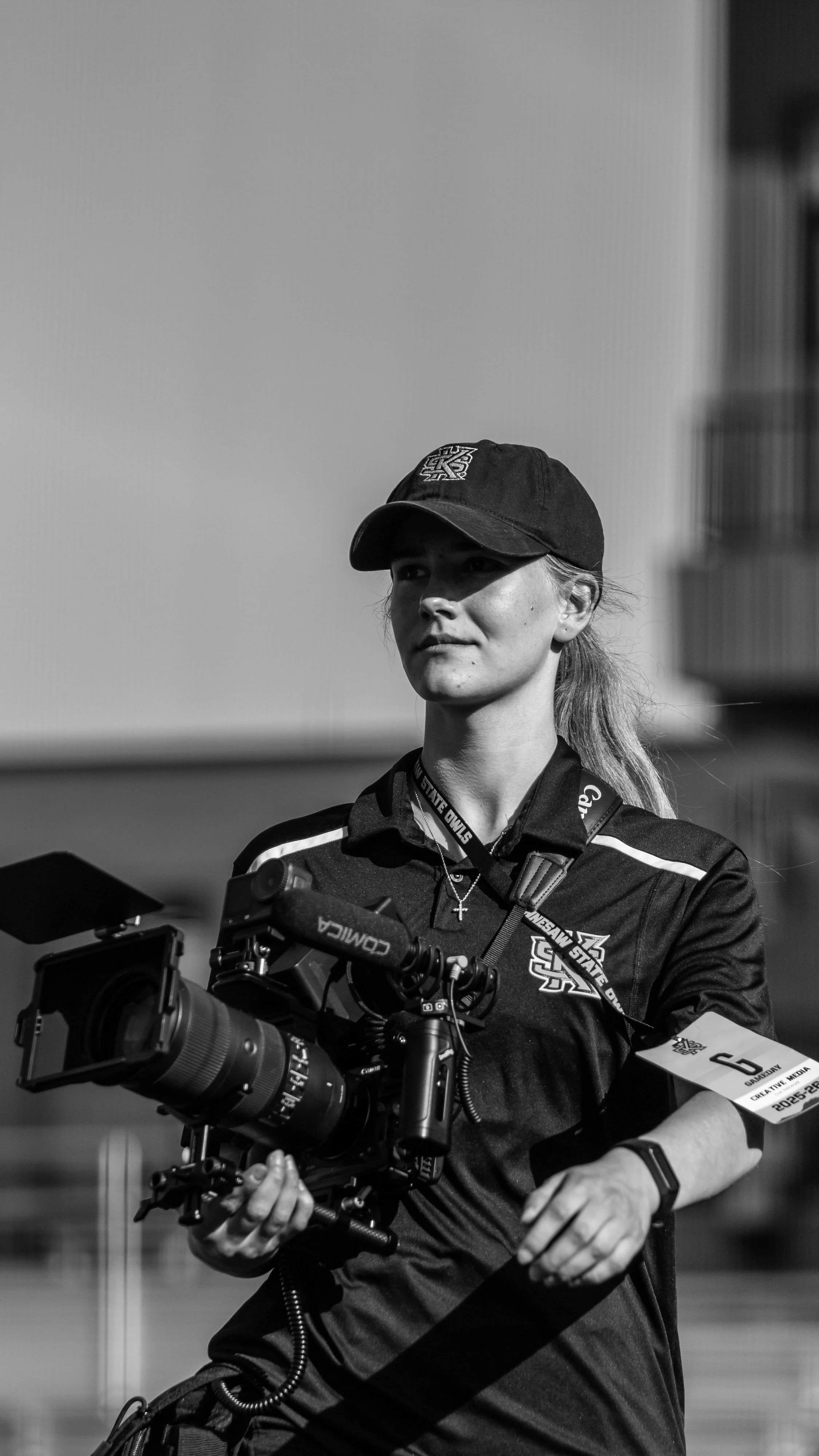A woman with light hair wearing a baseball cap and sports jersey, sitting with camera equipment, at an outdoor event.