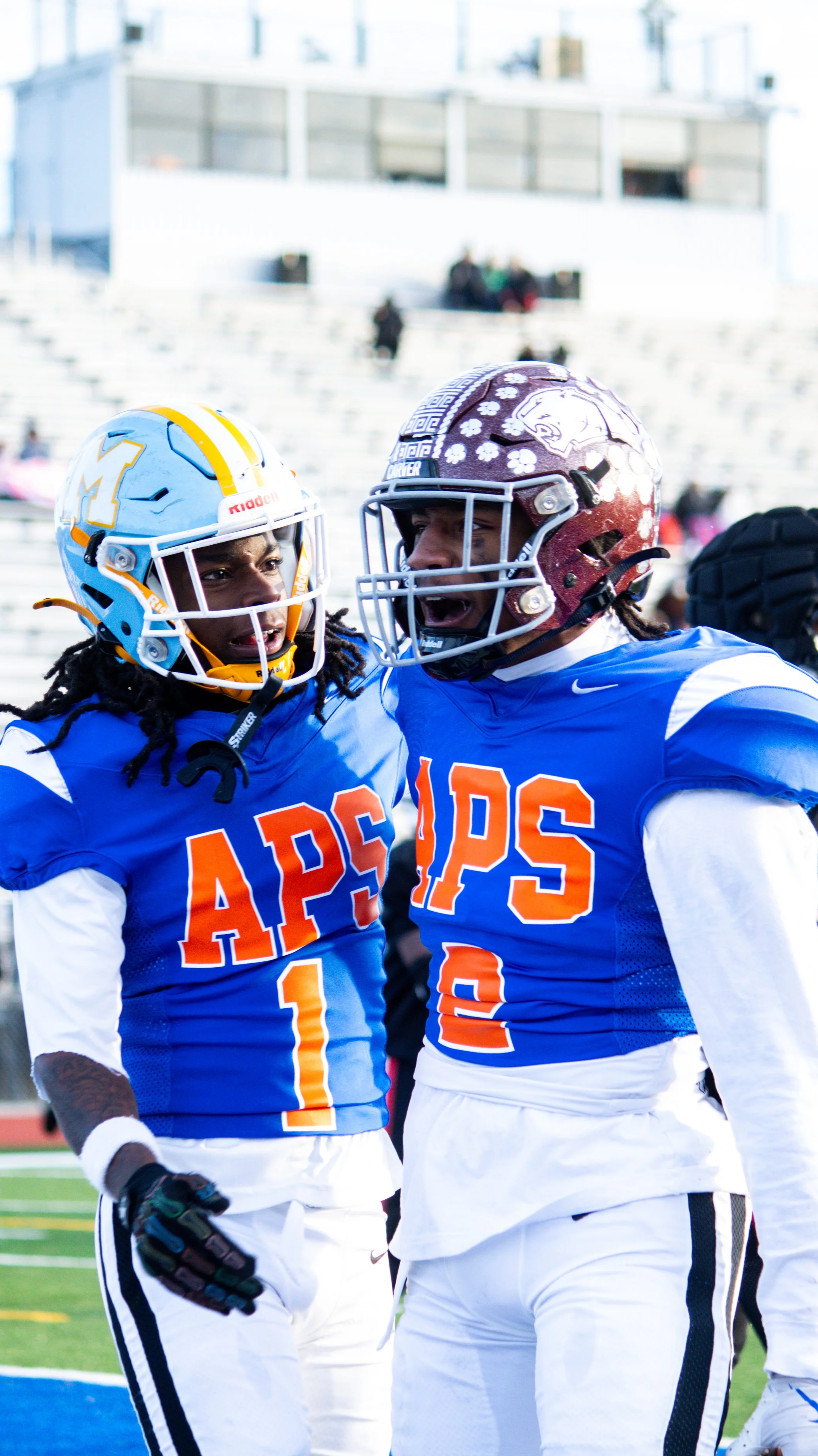 Two American football players wearing blue jerseys with 'APPS' written on them, helmets with Michigan and South Carolina logos, and white pants, standing and talking on a football field during a game.