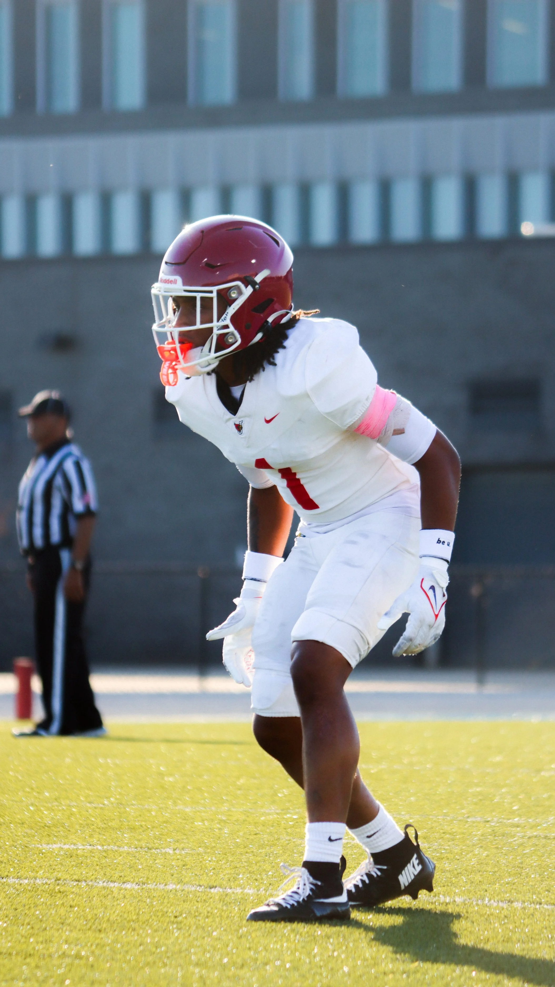 A football player in a white uniform with a maroon helmet and gloves, standing on a football field with a stadium background.
