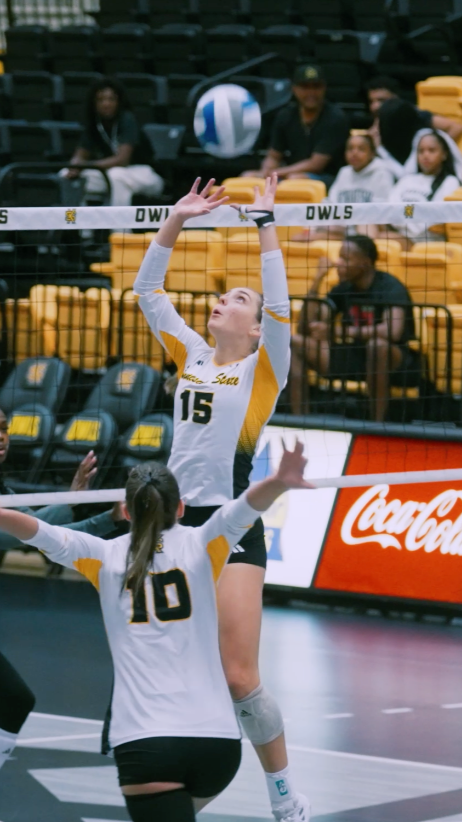 A volleyball player in a white and yellow jersey with the number 15 is jumping to hit the ball over the net, while another player in a white and black jersey with the number 10 is preparing to block or receive.