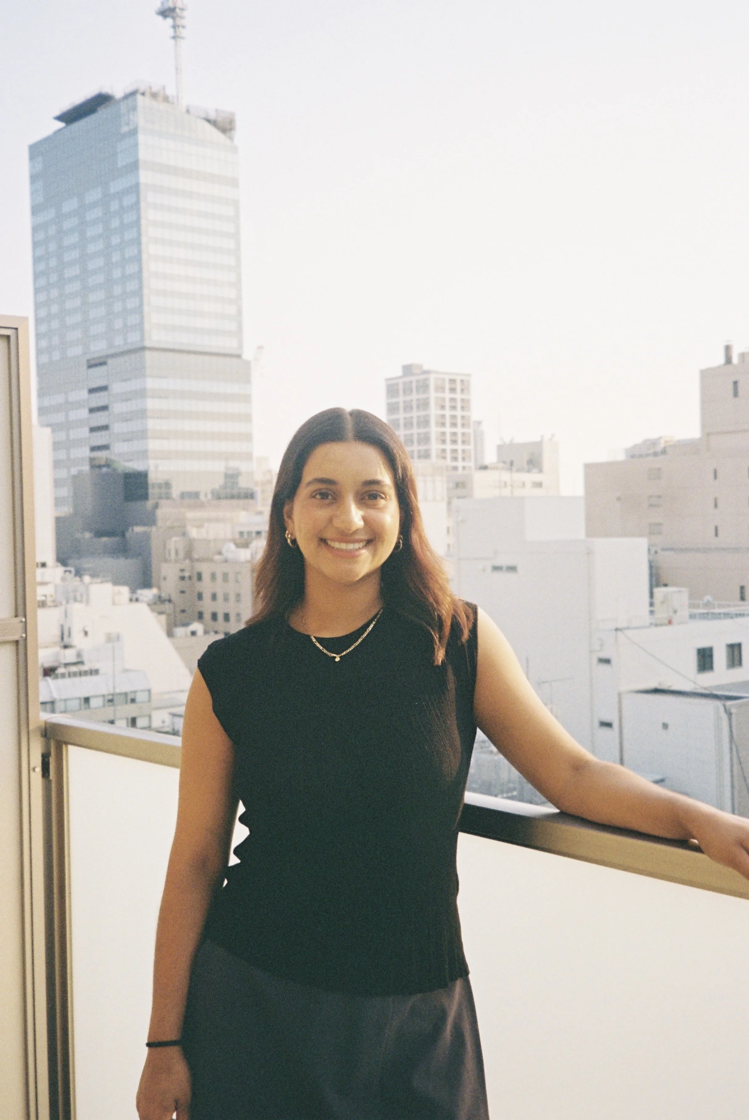 A woman with shoulder-length brown hair wearing a sleeveless black top and a necklace, smiling, standing on a balcony with city buildings in the background during daylight.