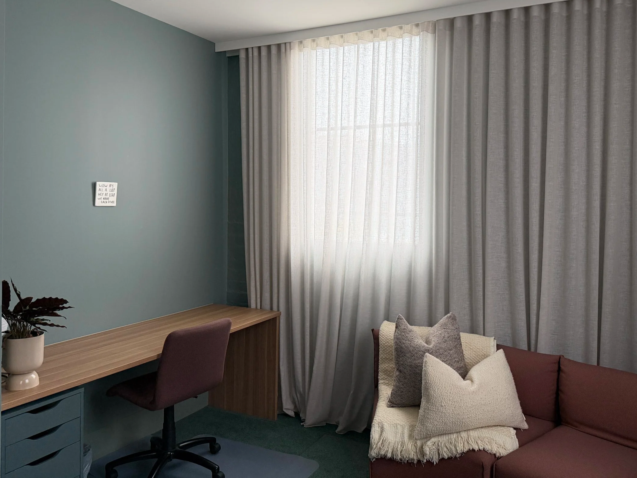 Modern living room corner with a blue wall, a wooden desk with a purple chair, a potted plant, and a large window with sheer curtains. A small sign hangs on the wall.