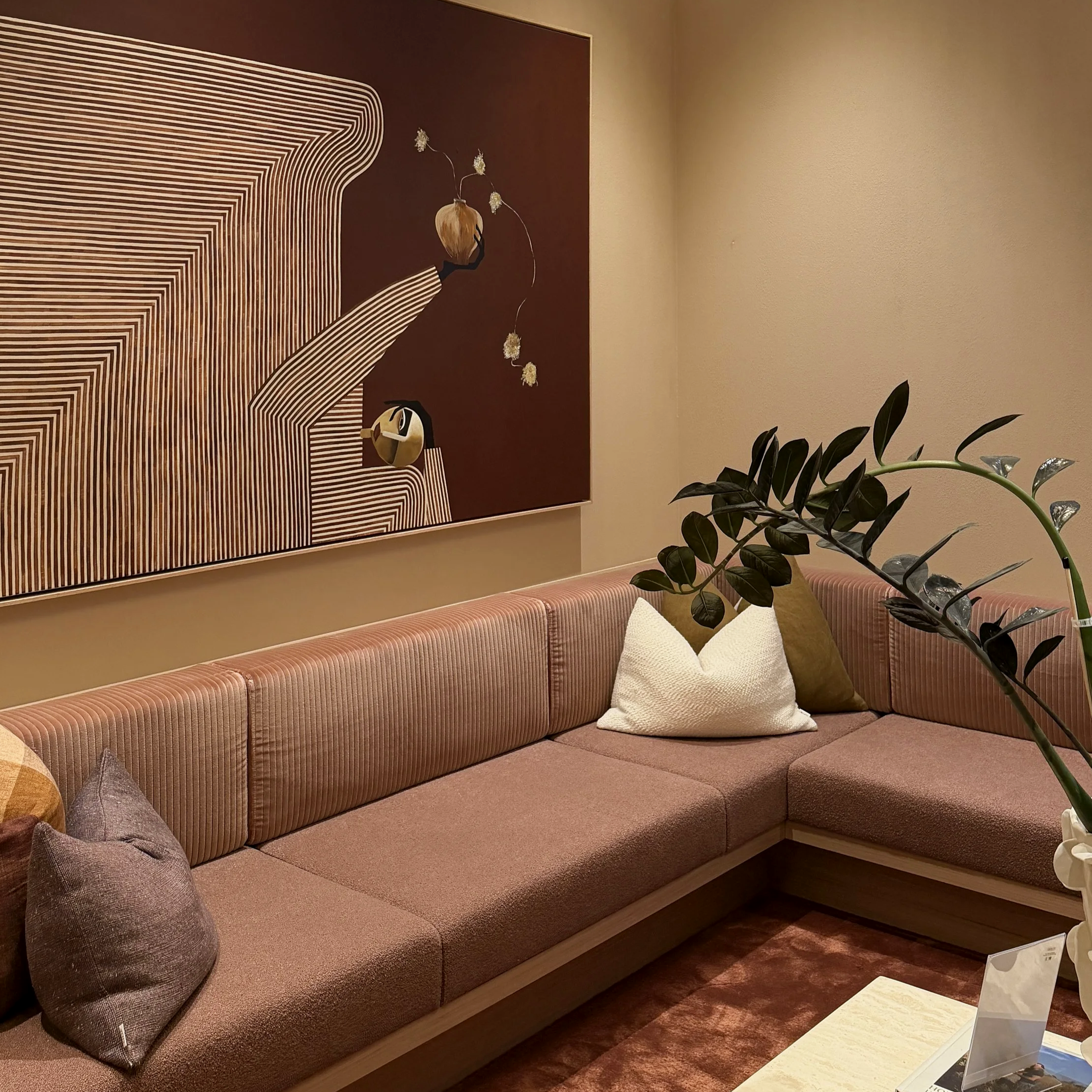 Living room with pink upholstered corner sofa, decorative pillows in beige and dark colors, a potted plant in the foreground, and a large abstract painting with brown, white, and beige geometric lines and floral accents on the wall.