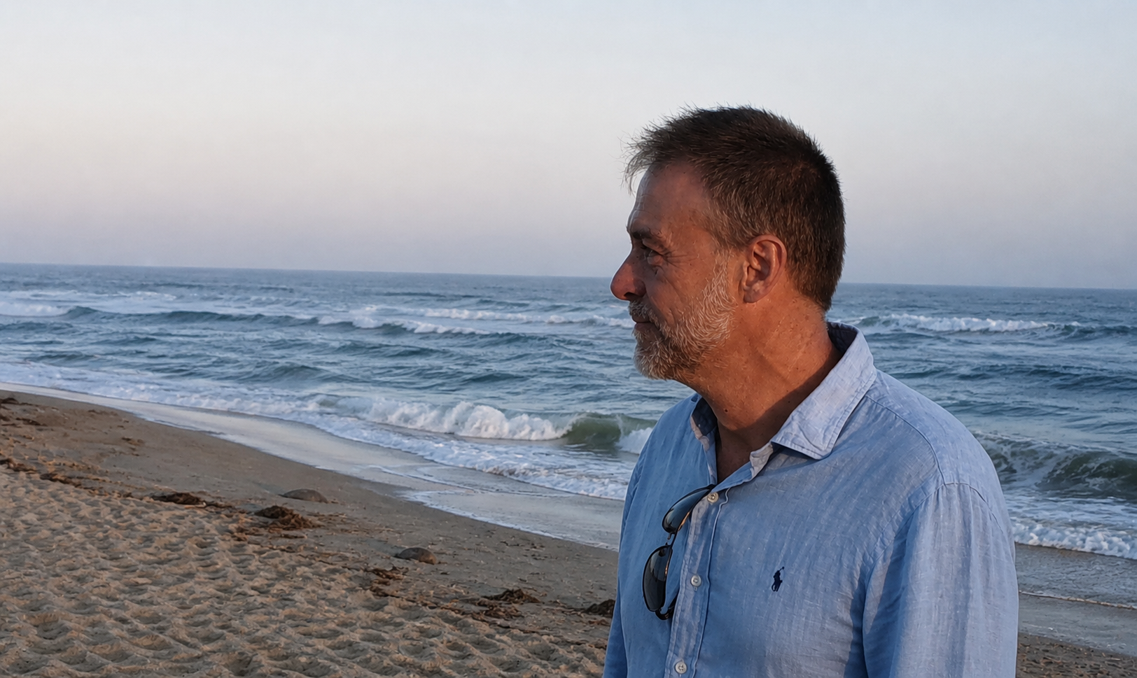 Man with short hair and beard wearing a light blue button-up shirt, standing on the beach near the ocean during sunset.