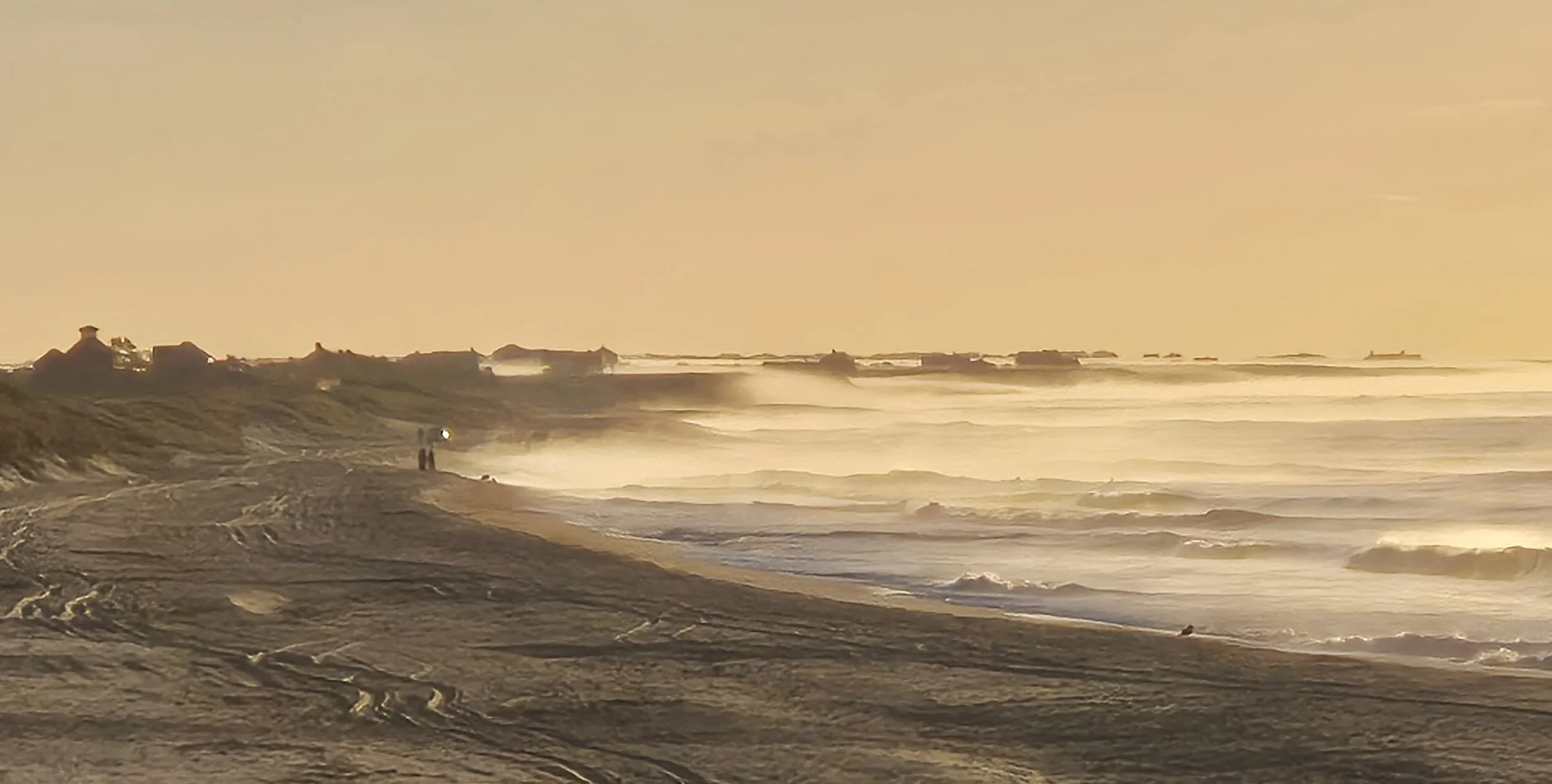 A beach at sunset with waves crashing on the shore and a few people walking along the beach.