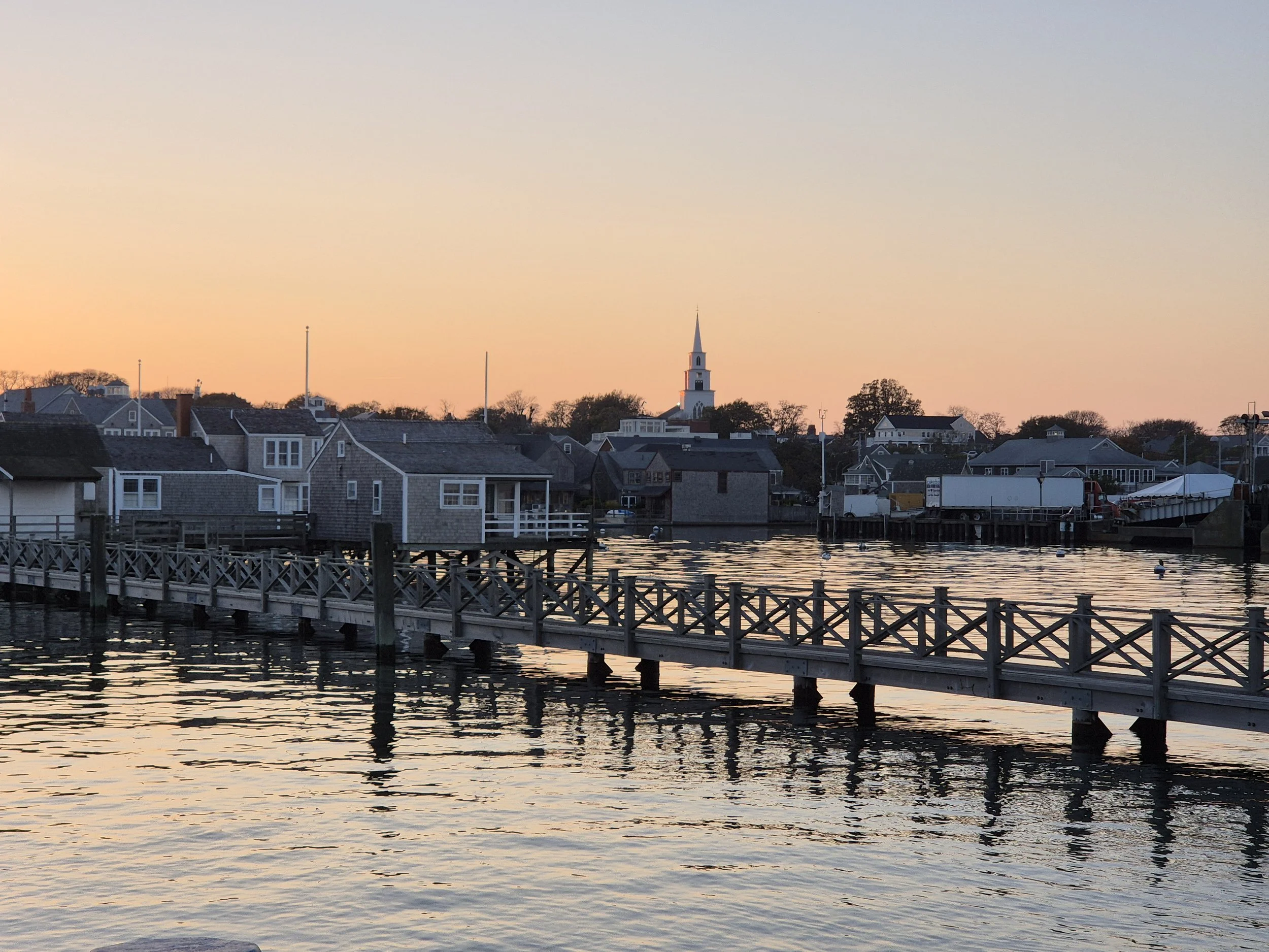 A peaceful seaside scene at dusk or dawn featuring a small town with houses, a church steeple, and a dock extending into calm water, with a pastel-colored sky overhead.