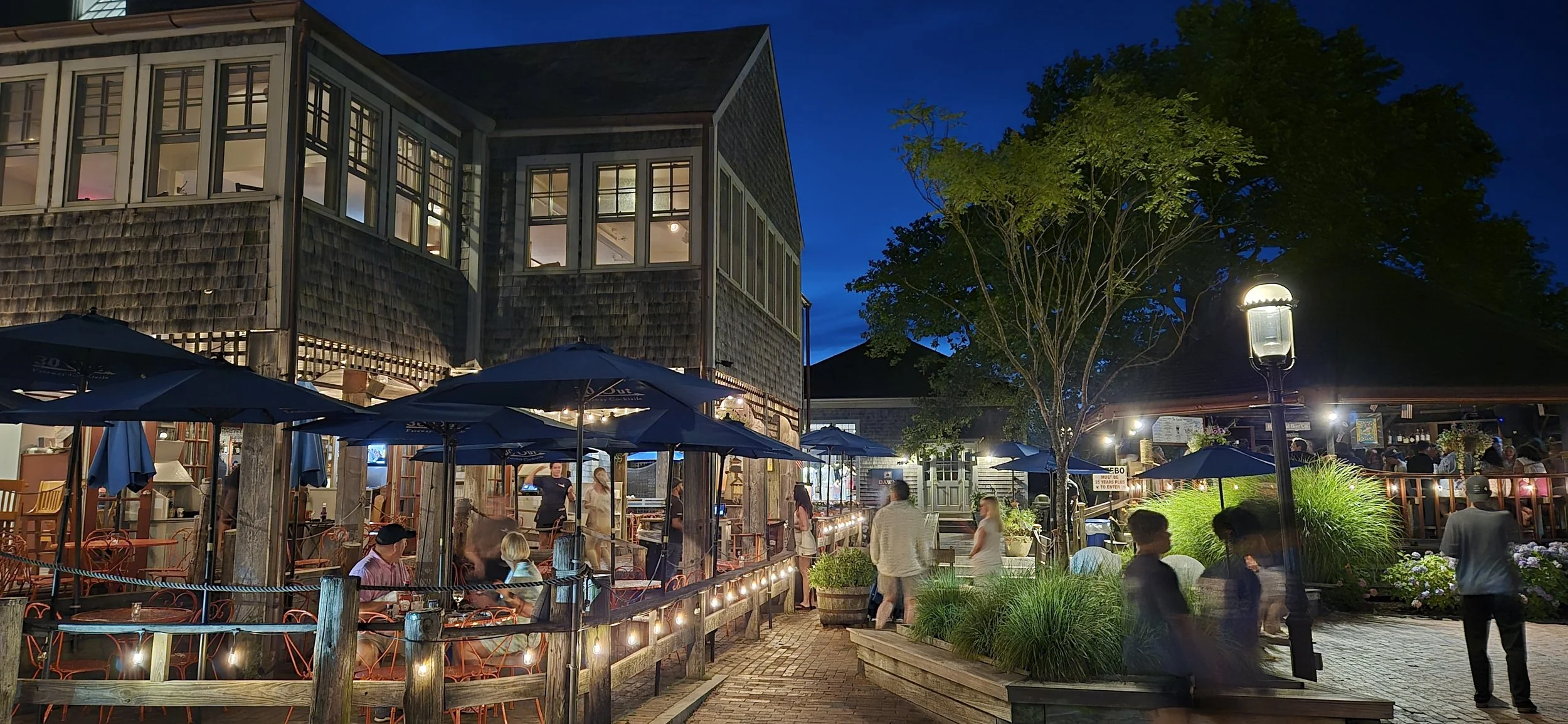 Outdoor restaurant patio at night with people dining, string lights, umbrellas, and a building with large windows and a tree.