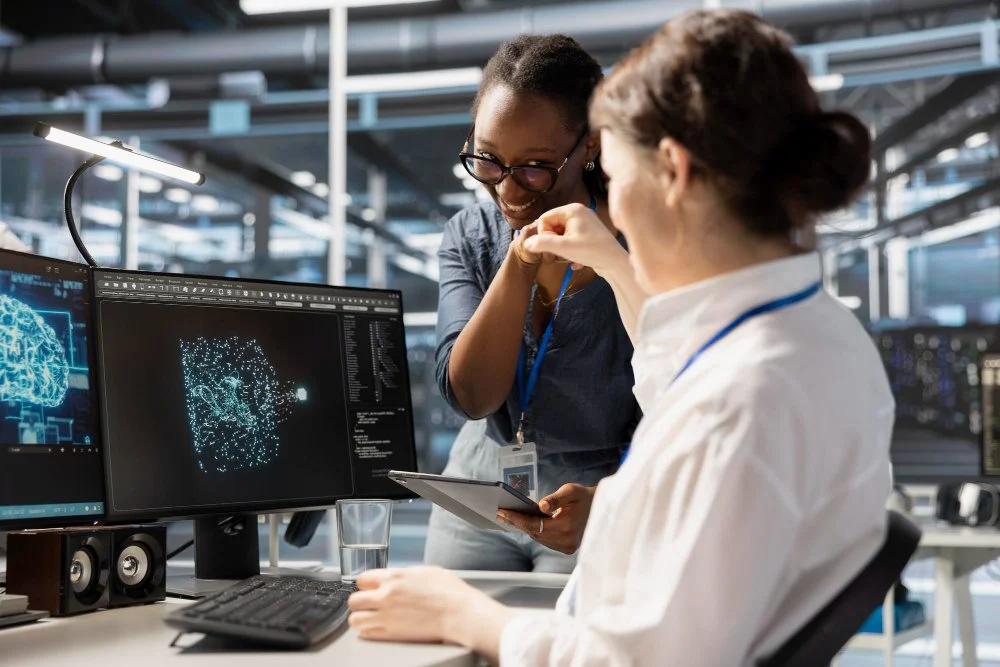 Deux femmes dans un laboratoire high-tech, l'une assise à un bureau avec deux écrans montrant des analyses de données, et l'autre debout partageant un moment de plaisir ensemble.