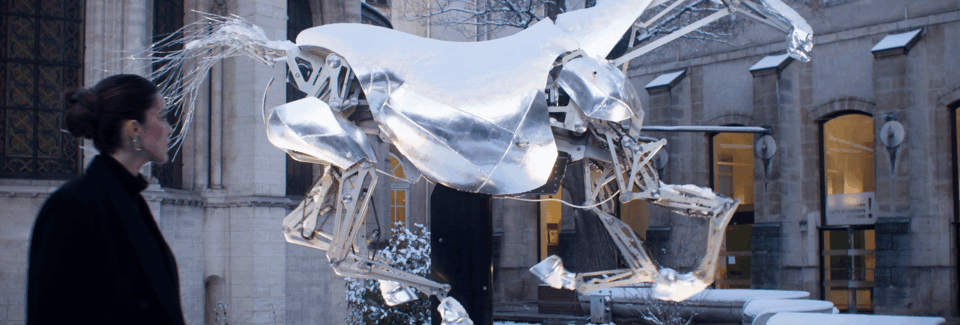 A woman observing a large metallic horse sculpture in an outdoor urban setting, with historic buildings in the background.
