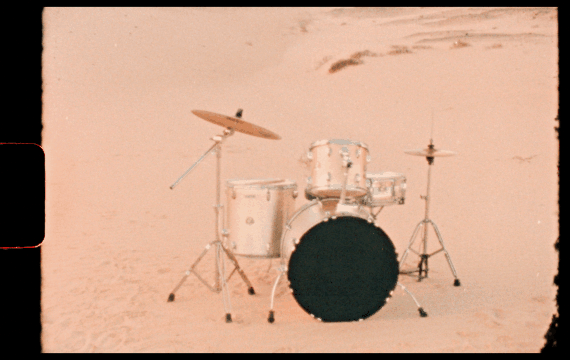A white drums set including a bass drum, snare drum, and hi-hat, set up on a sandy desert with dunes in the background.