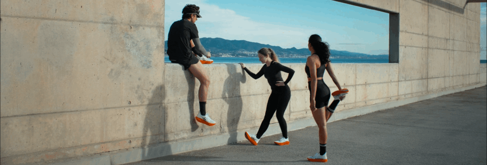Three women and one man in athletic clothing, conversation near a concrete wall with a large rectangular opening, body of water and mountains in the background.