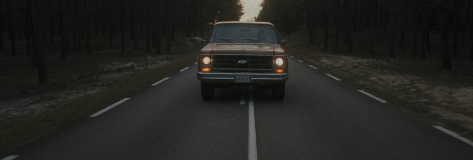 A classic pickup truck driving on a rural road through a forested area at sunset, the headlights are on.