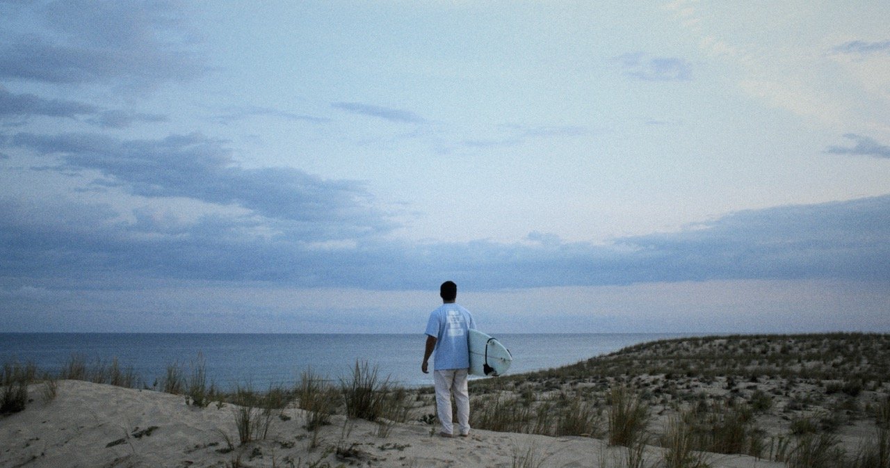 A man holding a surfboard standing on a sandy beach, overlooking the ocean during dawn or dusk with cloudy skies.
